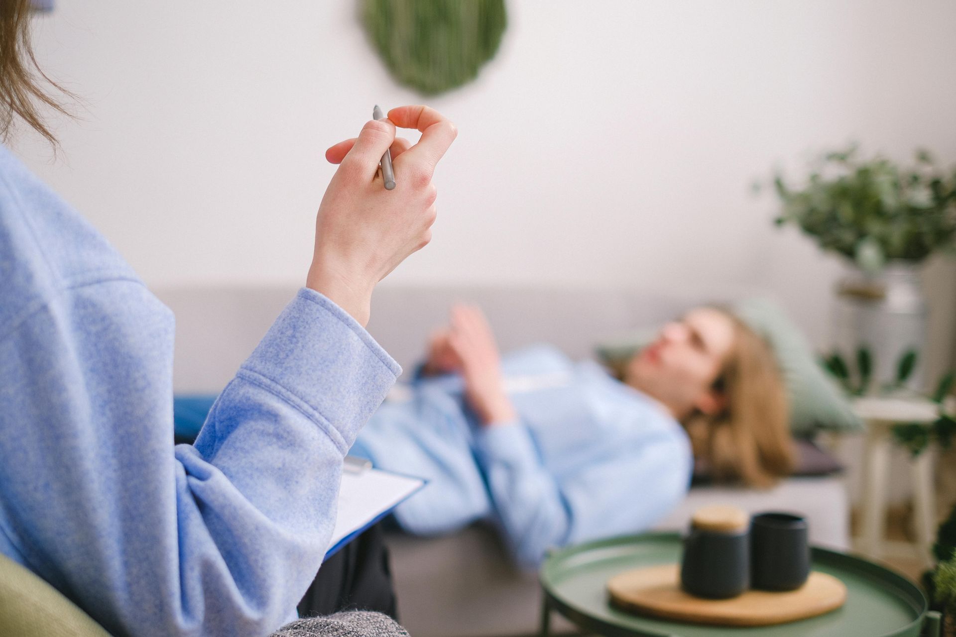 A person holds a pen and clipboard, facing another person who is sitting on a sofa during a counseling session.