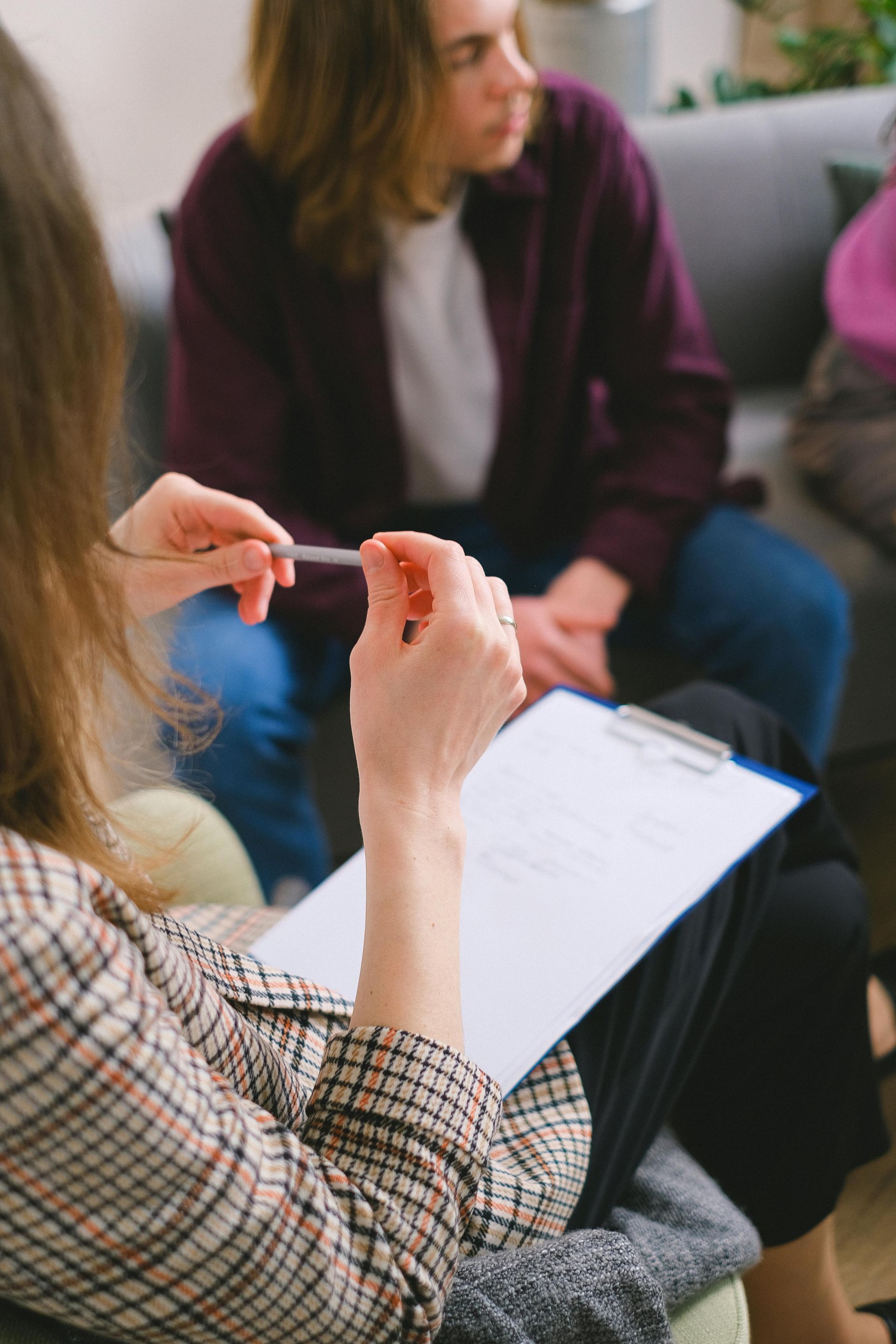 A person holds a pen and clipboard, facing another person who is sitting on a sofa during a counseling session.