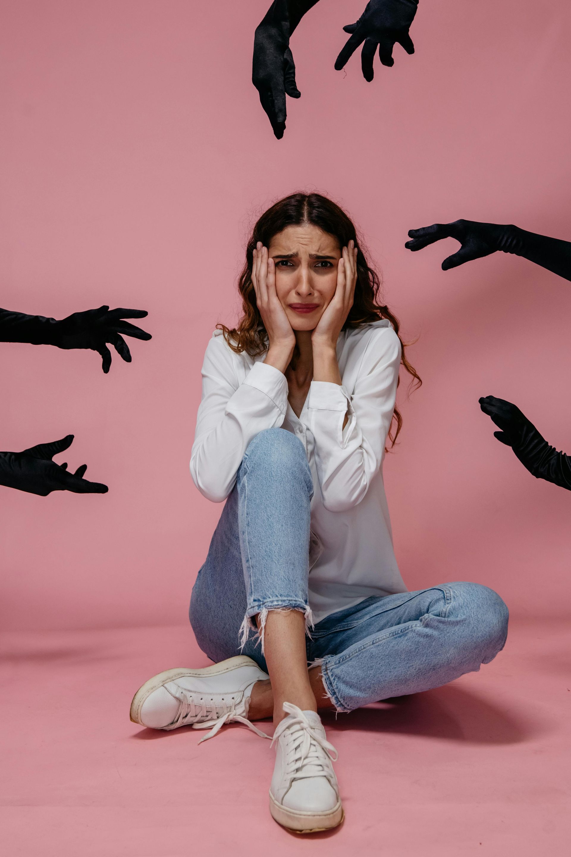 A person sitting on a pink floor looks distressed, holding their face as black-gloved hands reach toward them.