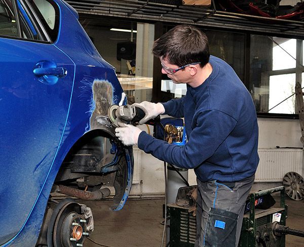 A technician in safety glasses and gloves uses an angle grinder to sand a damaged section of a blue car's rear fender.