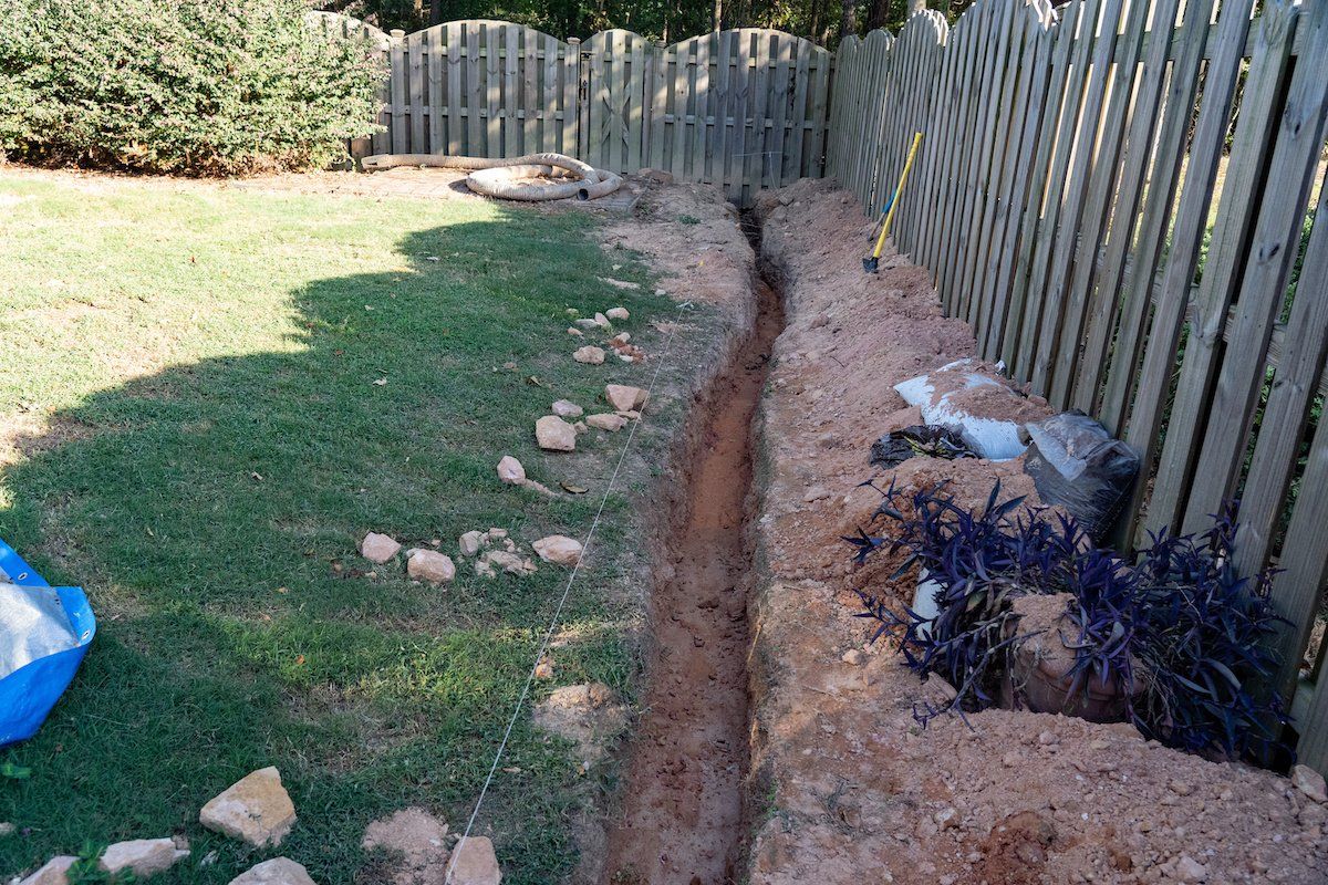 Backyard with a long trench beside a wooden fence and grass, exposing soil and rocks