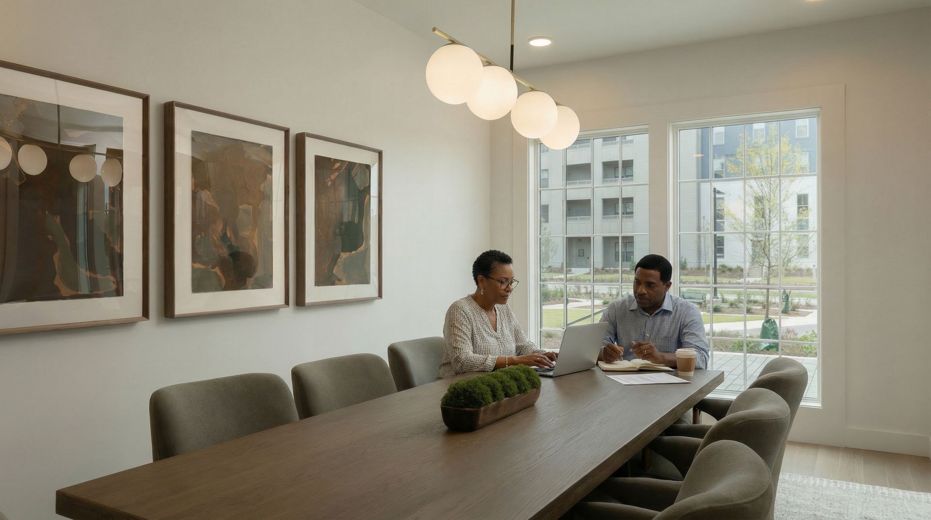 Two people working at a long table in a well-lit room with art and windows.