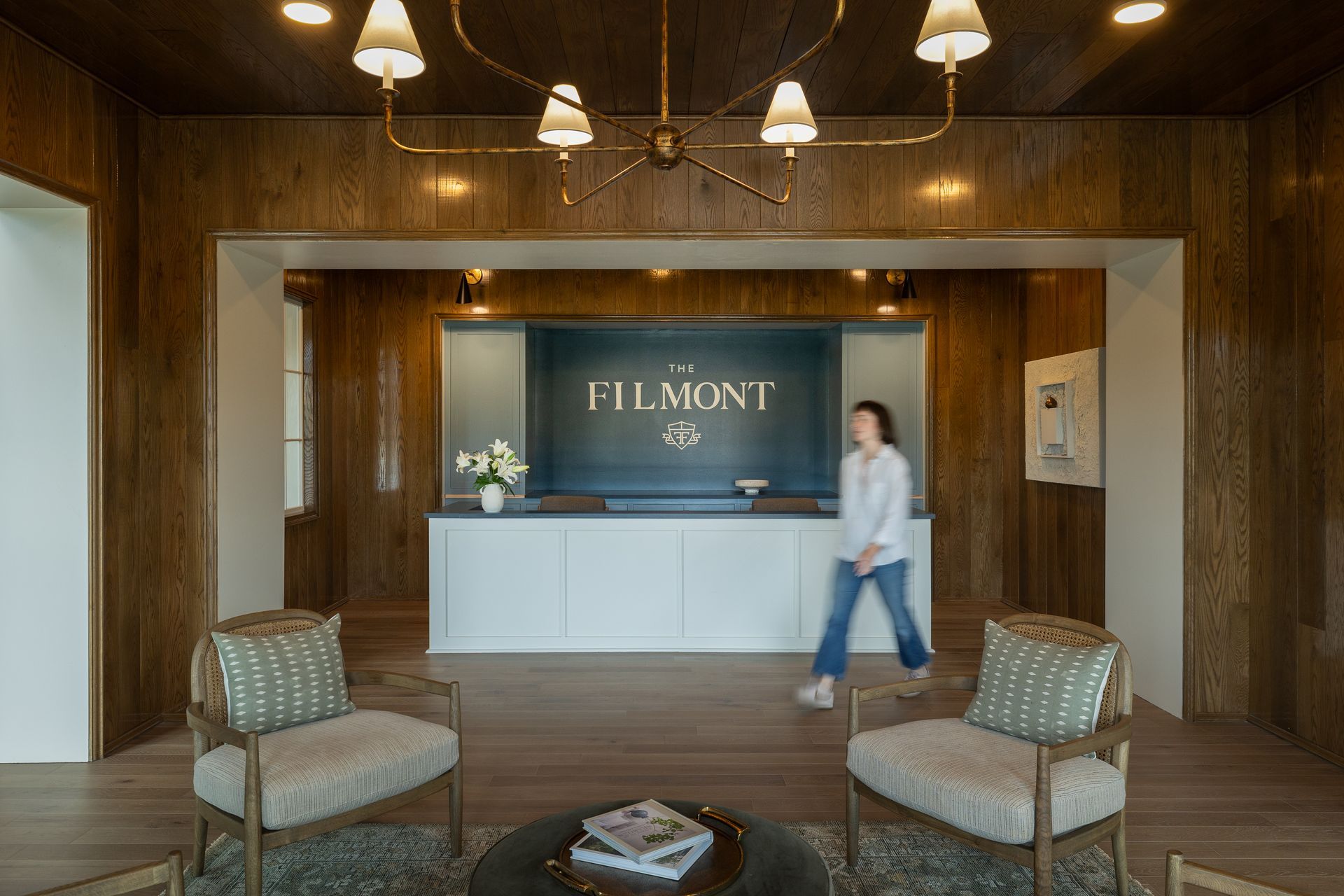 Elegant reception area with wood paneling, seating, and a person walking past the front desk, which has a logo that reads