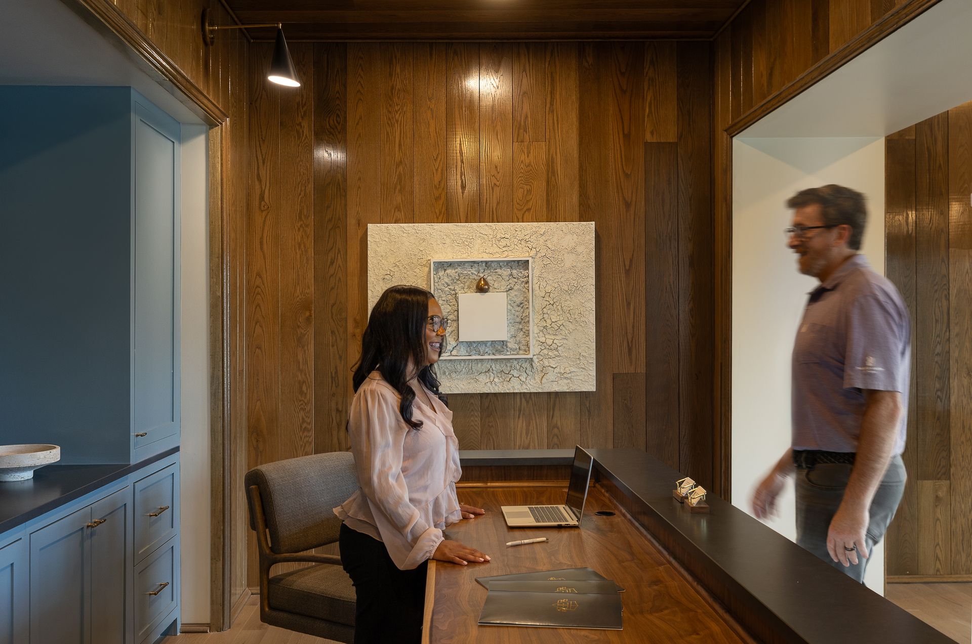 Woman at reception desk facing man approaching; wooden walls, art.