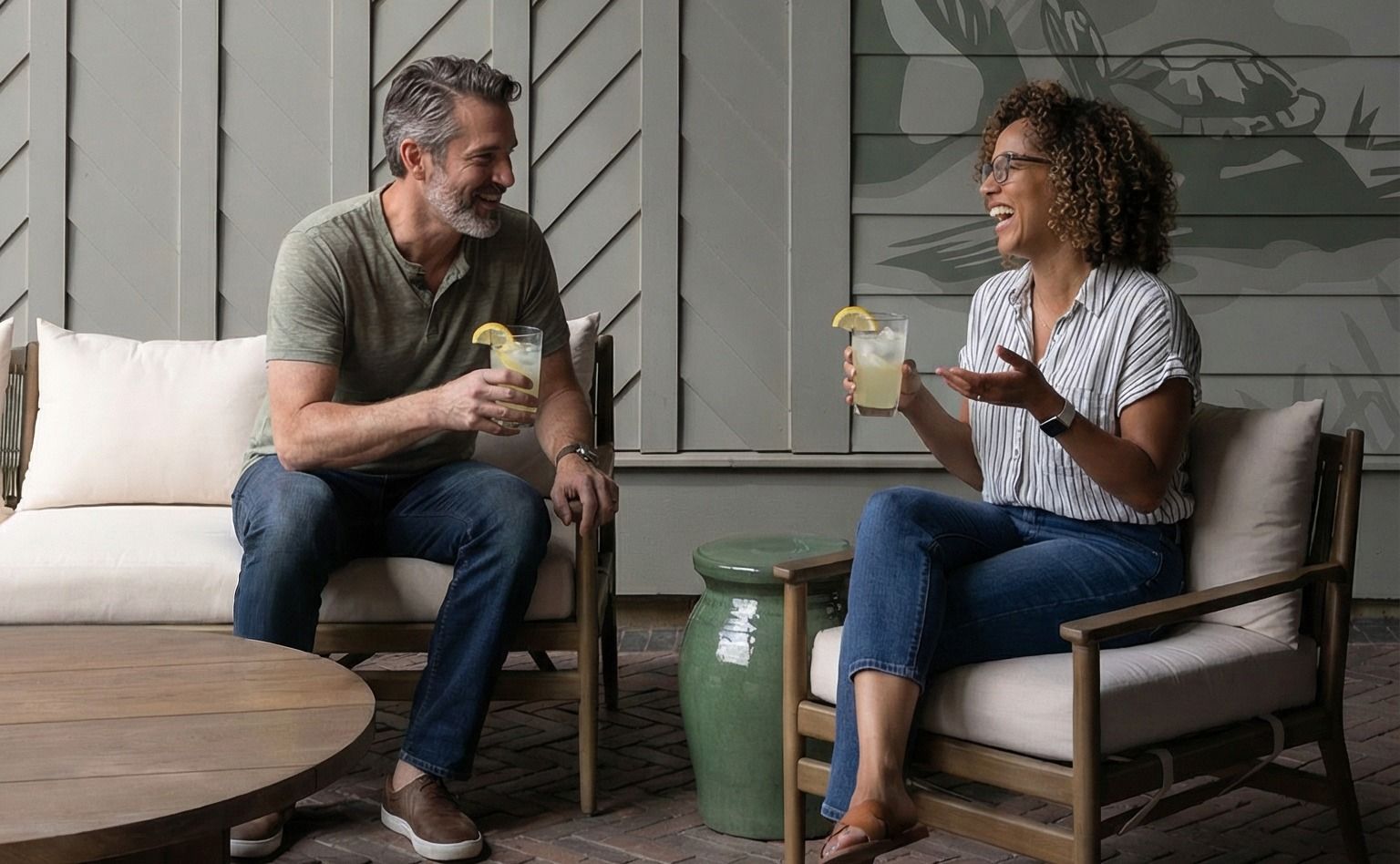 Couple seated on outdoor furniture, toasting with drinks. Gray wall with mural backdrop.