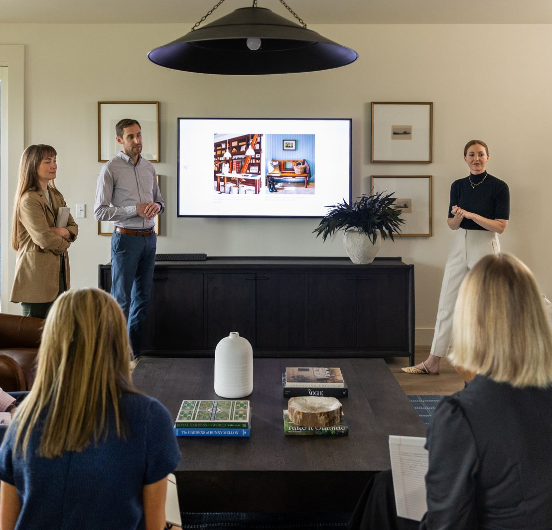 Two people working at a long table in a well-lit room with art and windows.