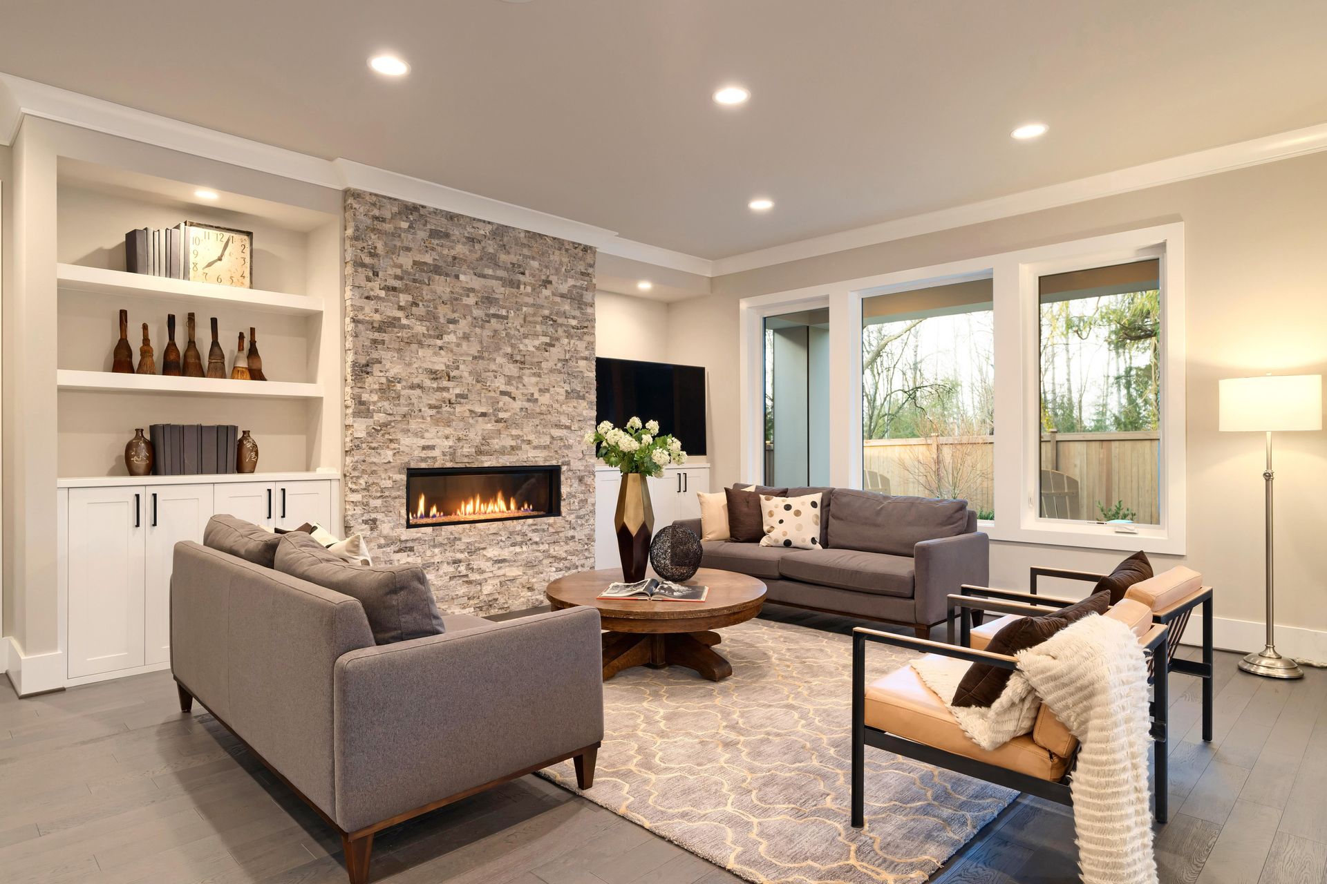 Cozy living room with a stone fireplace, gray sofas, a wooden coffee table, and a rug, alongside a large window.