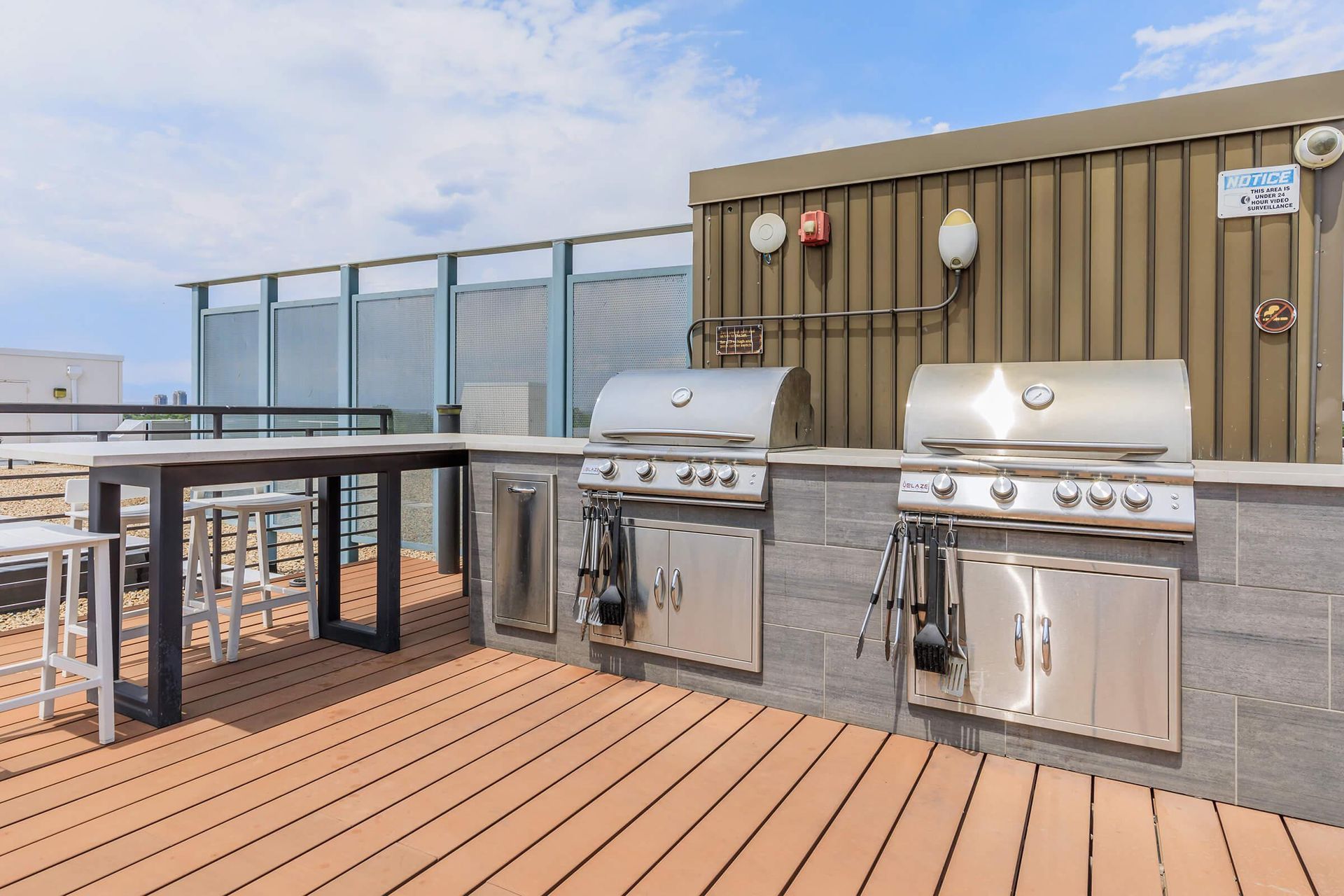 Rooftop grilling area with stainless steel grills, a table with stools, and a blue sky.