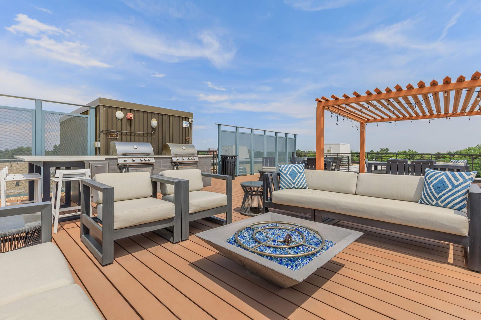 Rooftop deck with outdoor furniture, fire pit, grill, and pergola against a blue sky.