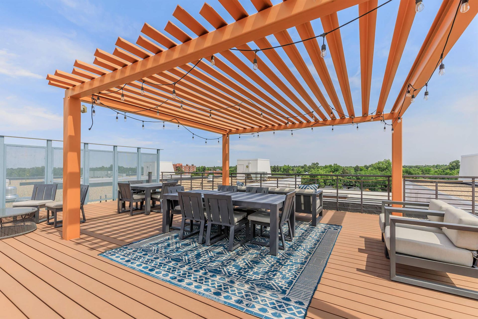 Rooftop patio with orange pergola, tables, chairs, blue rug, and string lights, overlooking a city.