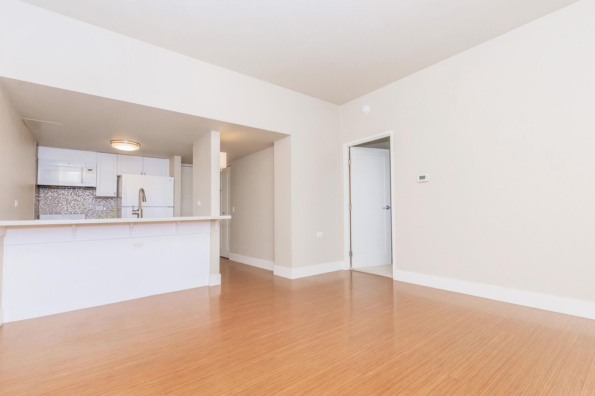 Empty apartment interior with hardwood floors and white walls; kitchen visible in the background.