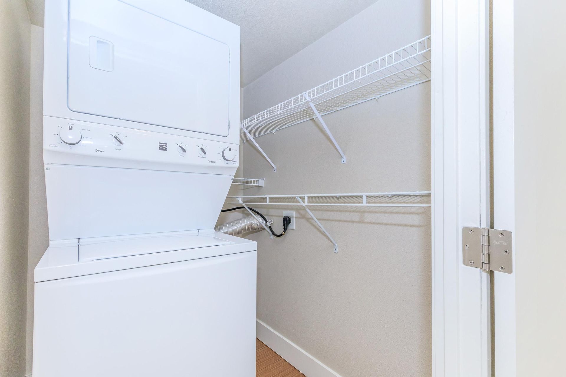 Stackable white washer and dryer in a small laundry closet with wire shelving.