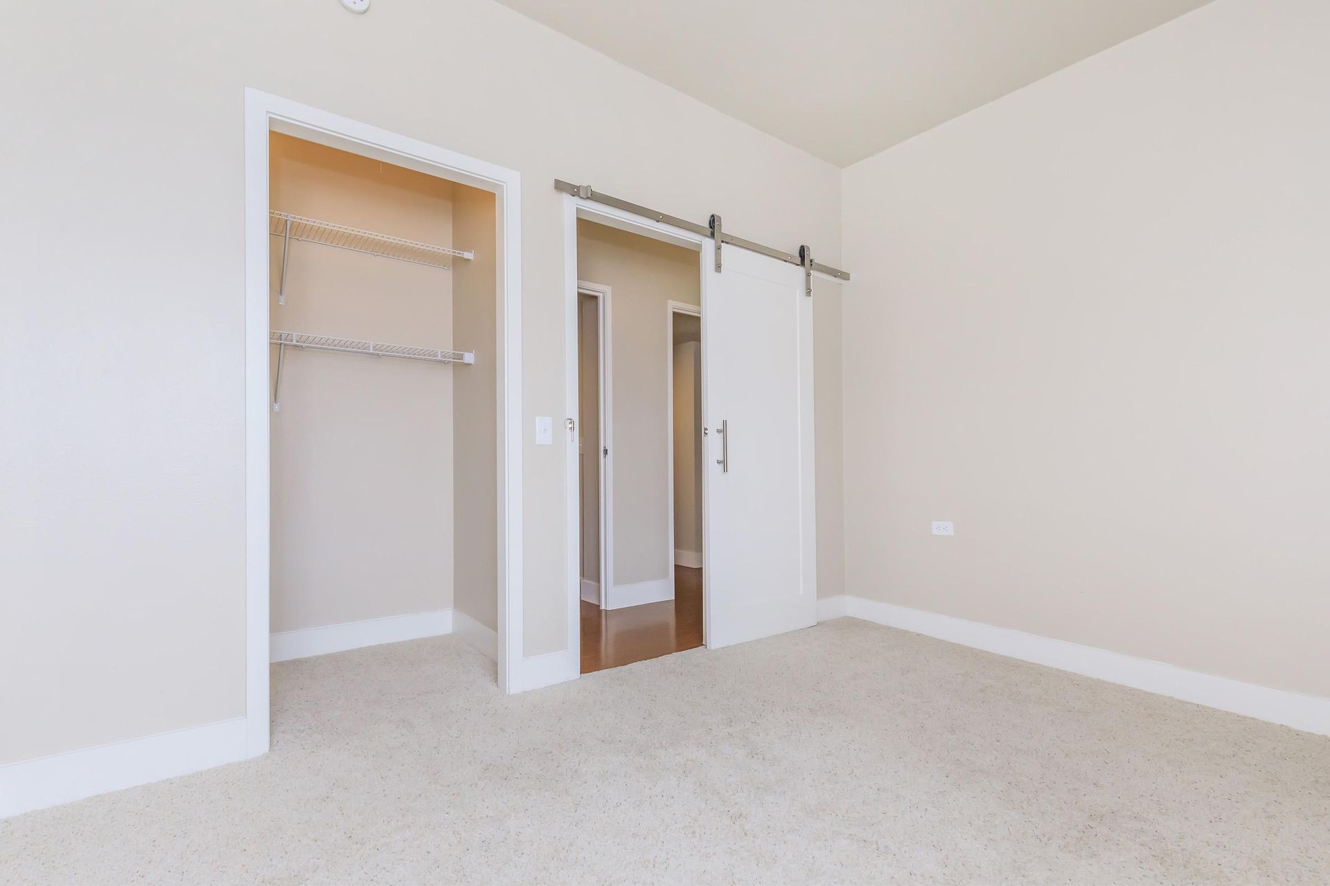 Empty bedroom with open closet and a sliding barn door leading to a hallway; cream walls, carpet.