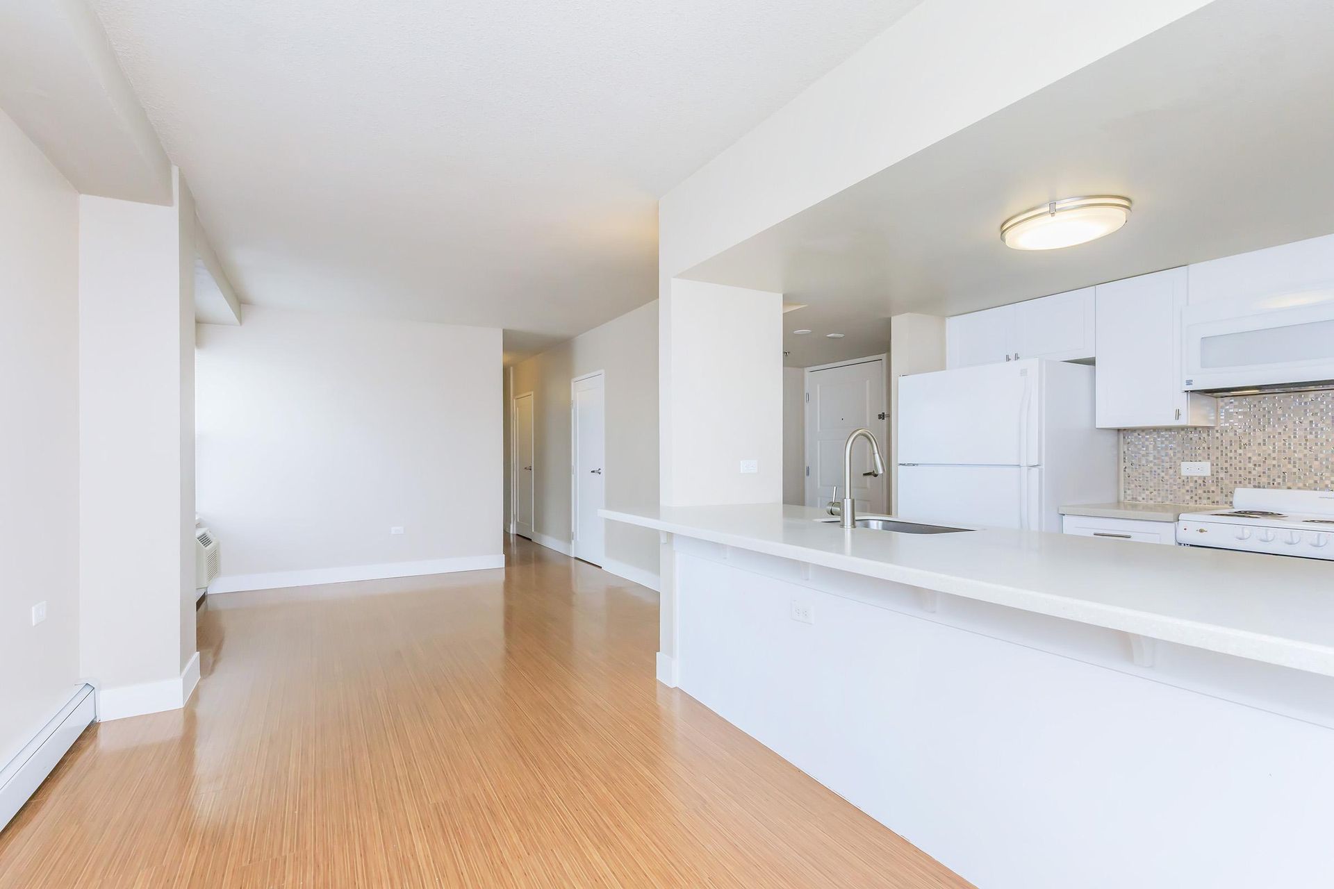 Bright, empty apartment with hardwood floors and white kitchen with a breakfast bar.