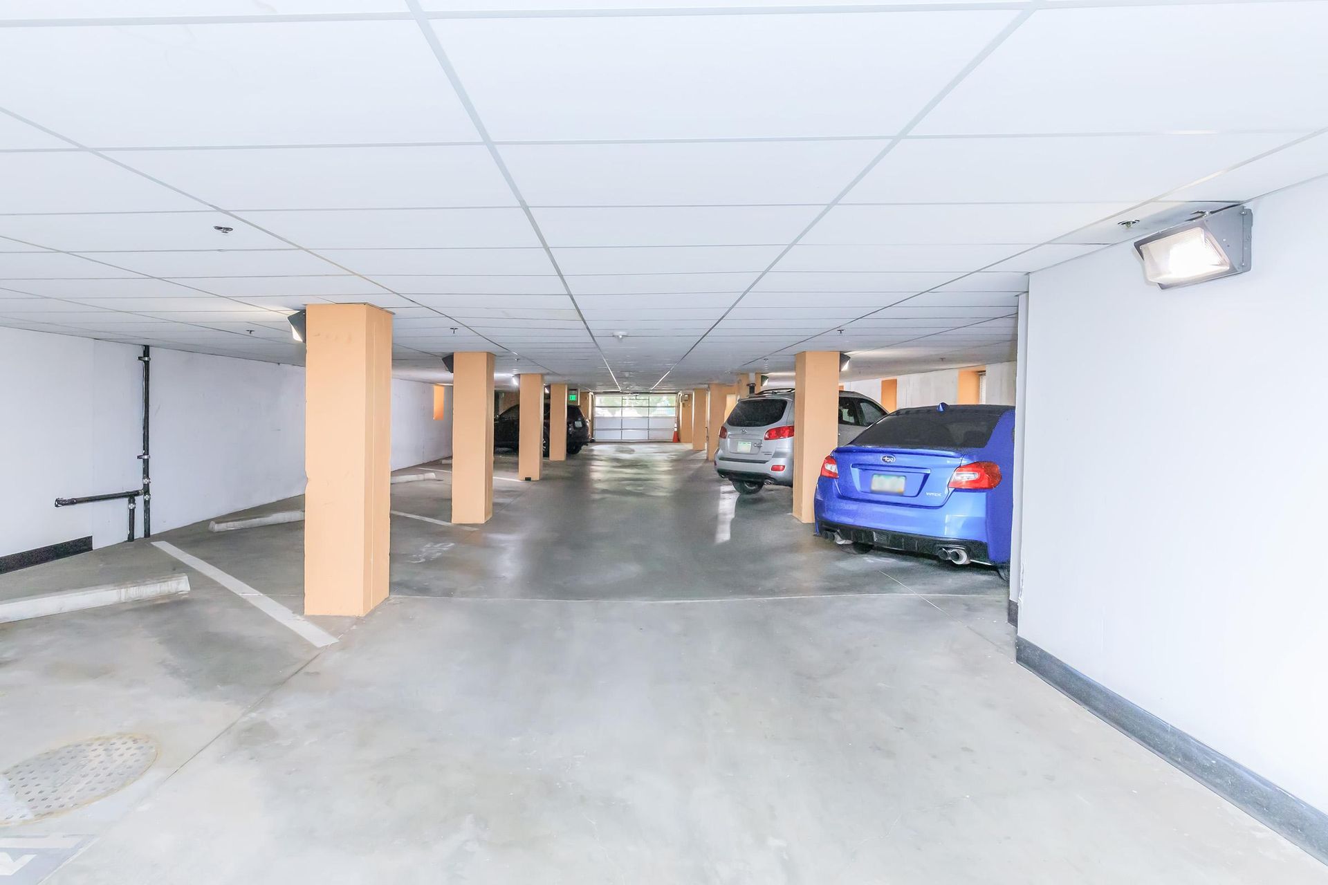 Underground parking garage with concrete floor, pillars, and parked cars. White walls and fluorescent lighting.
