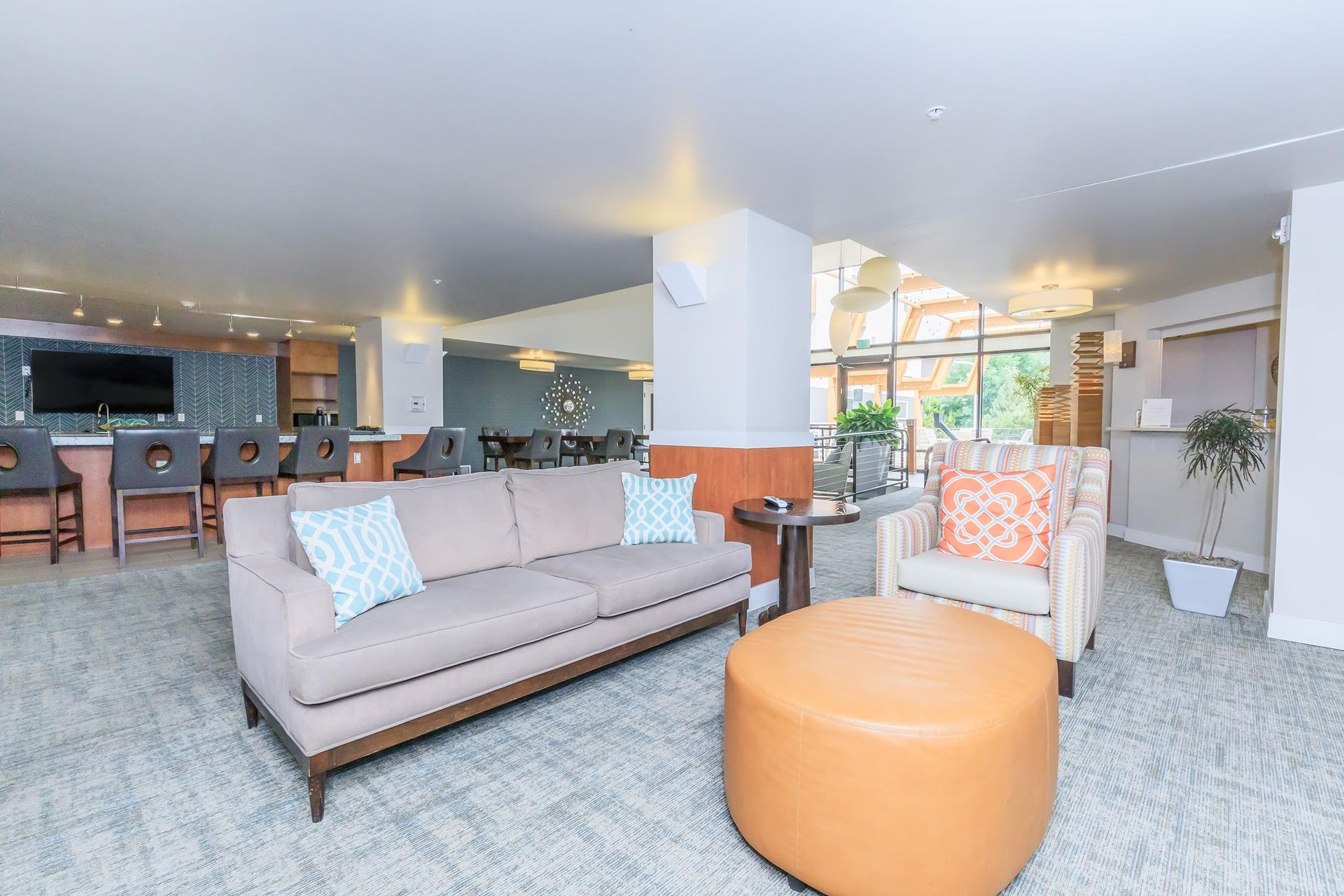 Lounge area with tan sofa, orange ottoman, patterned armchair, and bar in the background.