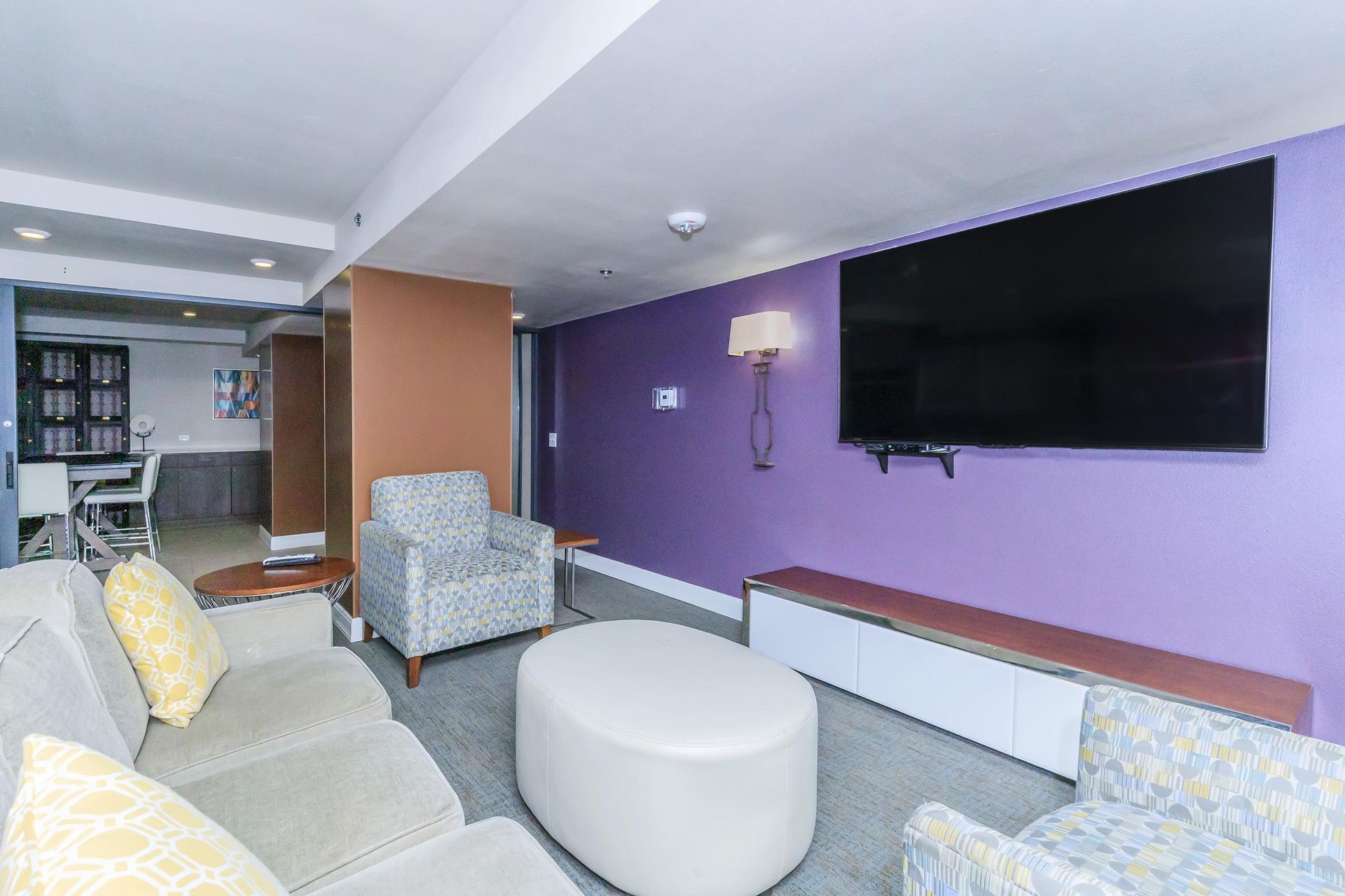 Living room with purple wall, large TV, white ottoman, and two armchairs.