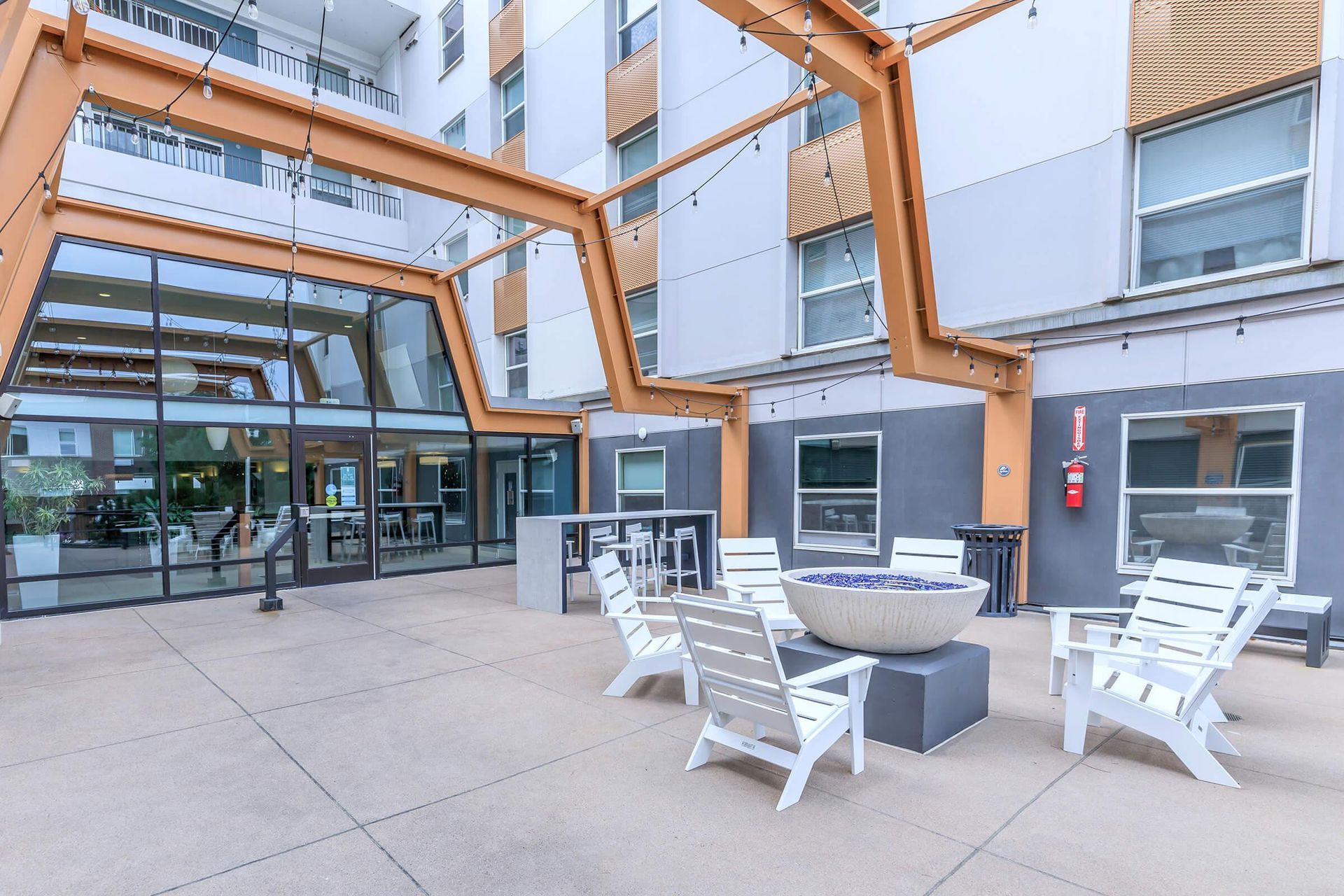 Outdoor patio with white chairs, a fire pit, and a glass entrance to a building.