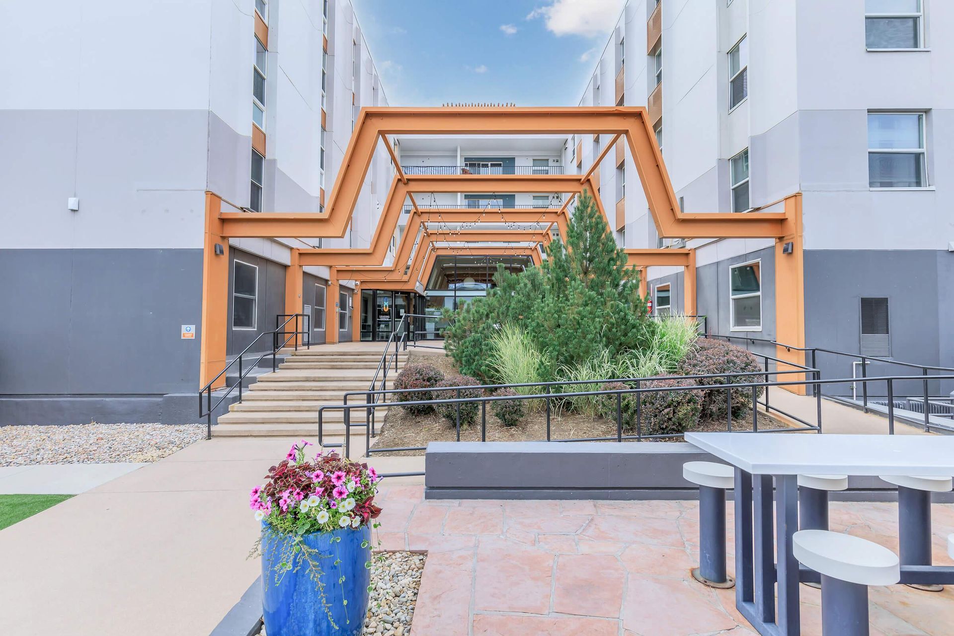 Entrance to a modern building with orange metalwork, stairs, a ramp, and outdoor seating.