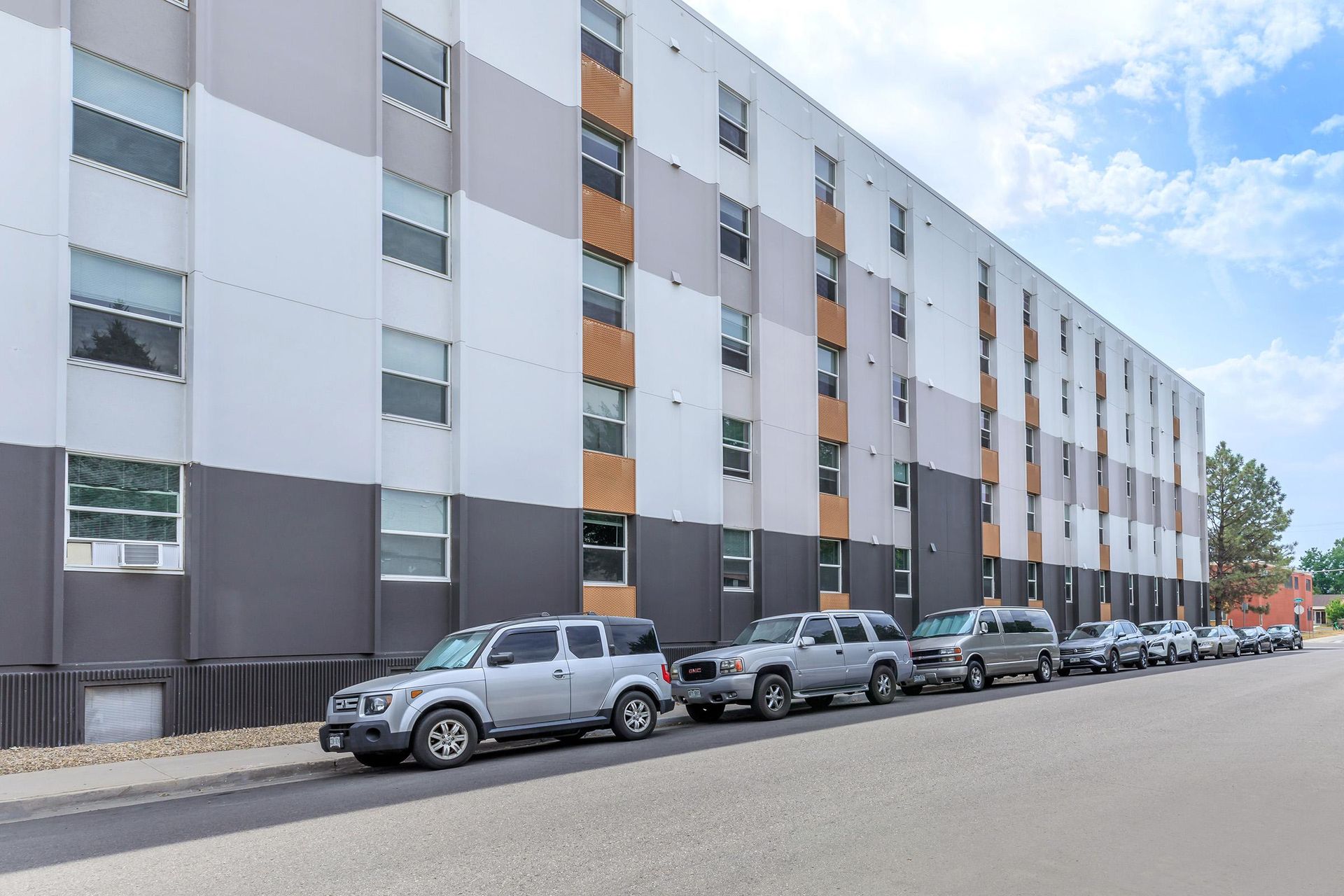 Apartment building exterior, four-story, with parked vehicles on street. Building has gray, white, and orange panels. Blue sky.
