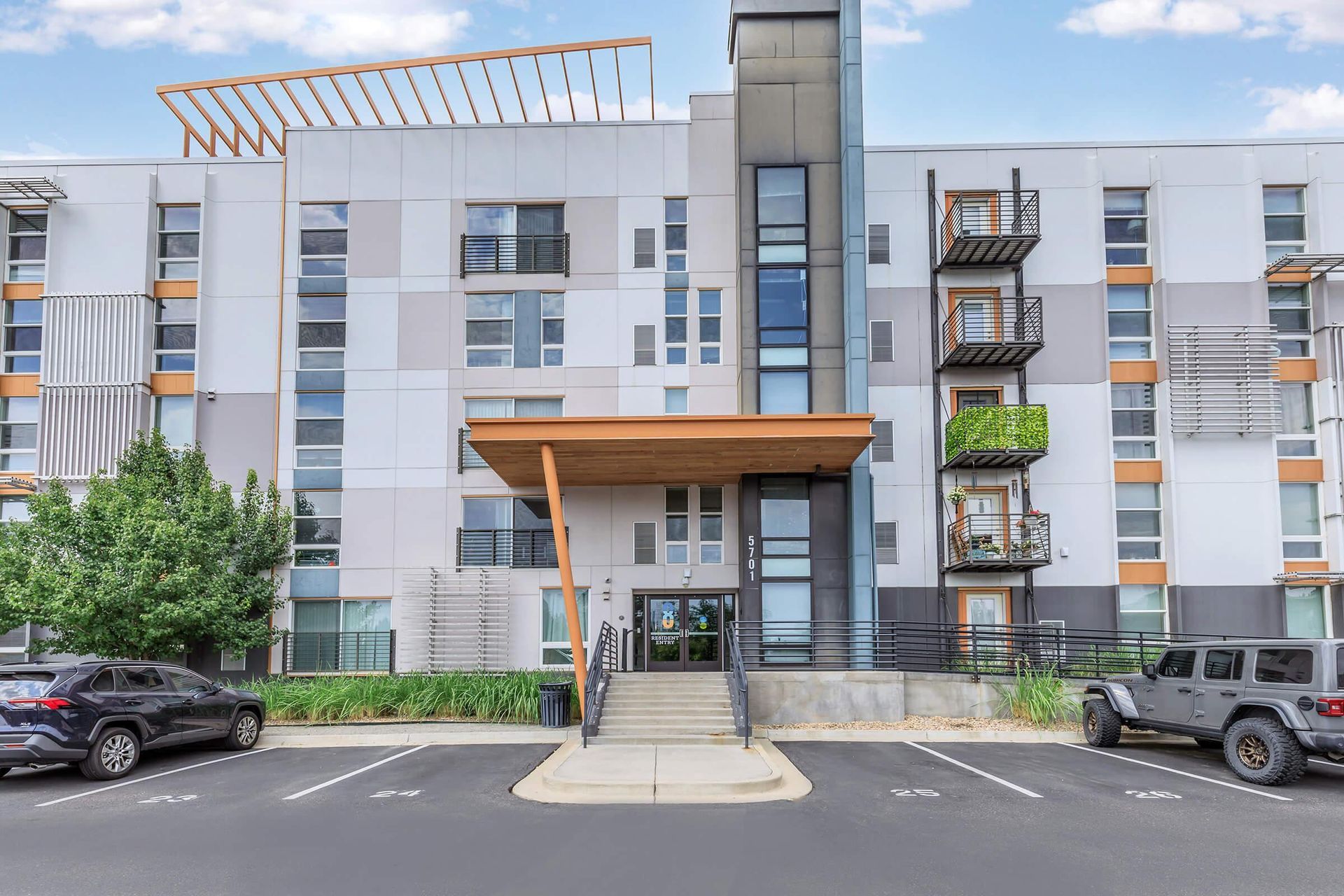 Modern apartment building exterior with parking. Gray and white facade, brown canopy, stairs to entrance.