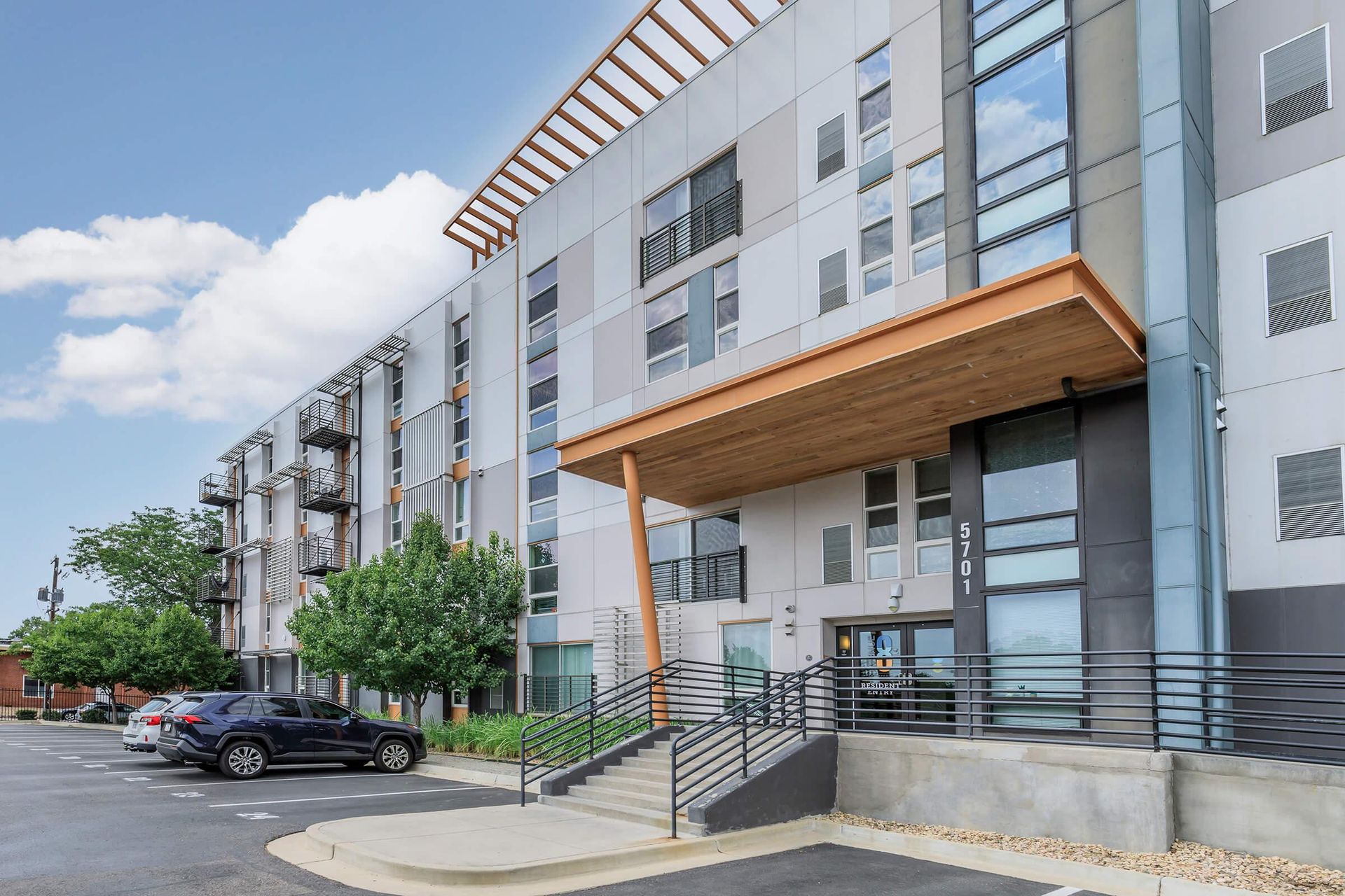 Modern apartment building with a wooden canopy over the entrance, and stairs. Cars parked in front.