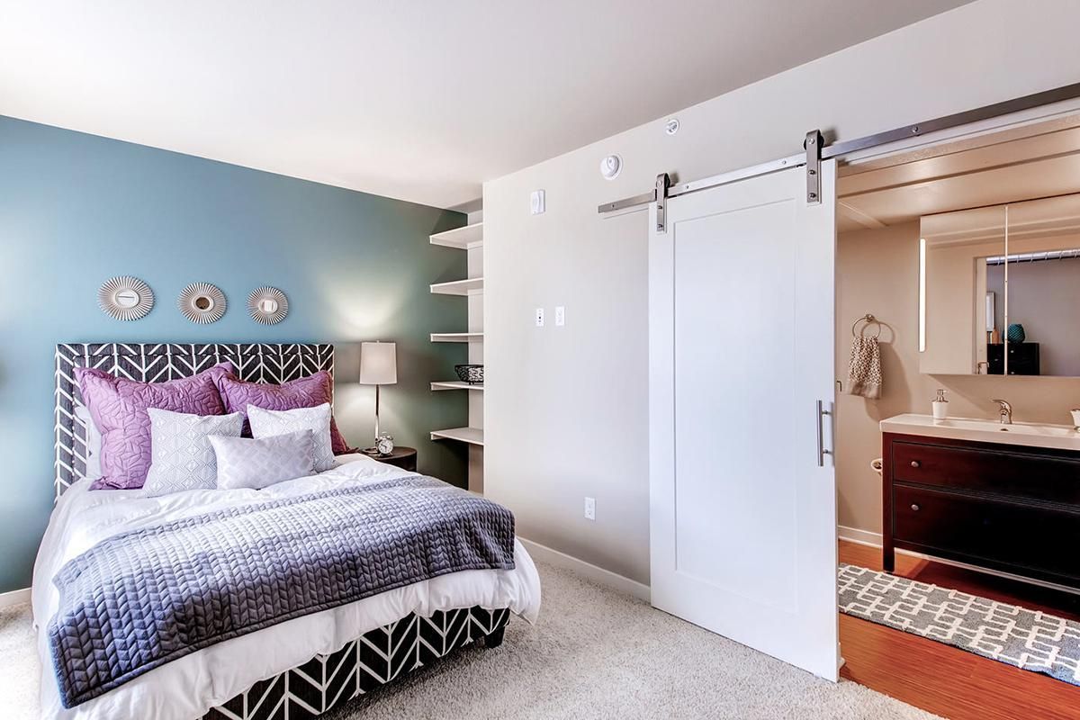 Bedroom with blue wall, bed with purple pillows, open white sliding door to bathroom.