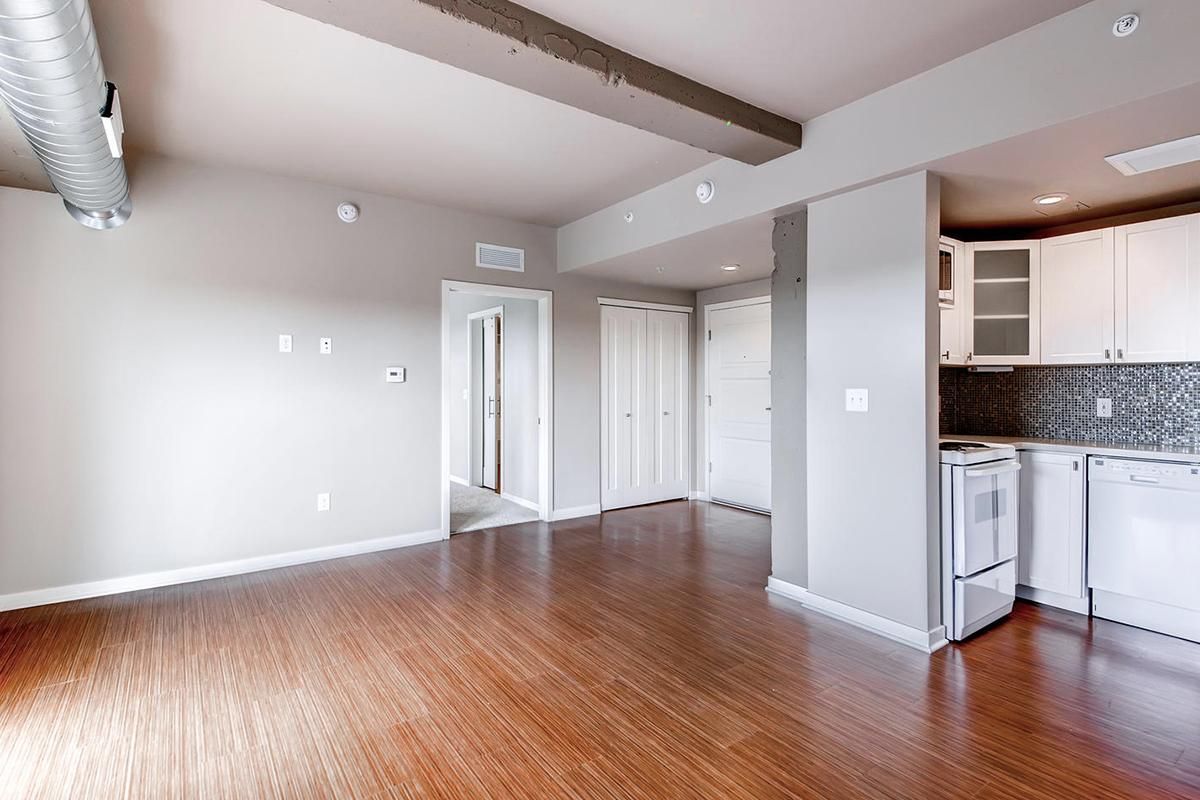 Empty living space with hardwood floors, white walls, and a small kitchen area.