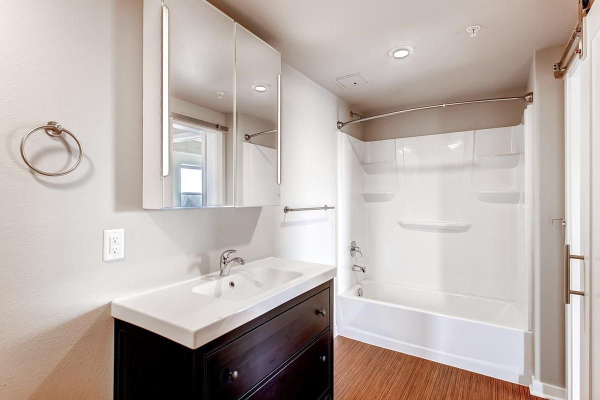 Bathroom with white walls, vanity, and shower/tub combo. Wooden floor.