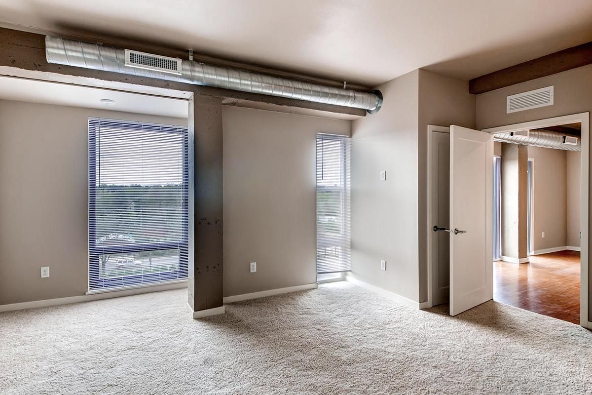 Empty room with neutral walls, carpet, and windows with blinds; a large metal duct runs along the ceiling.