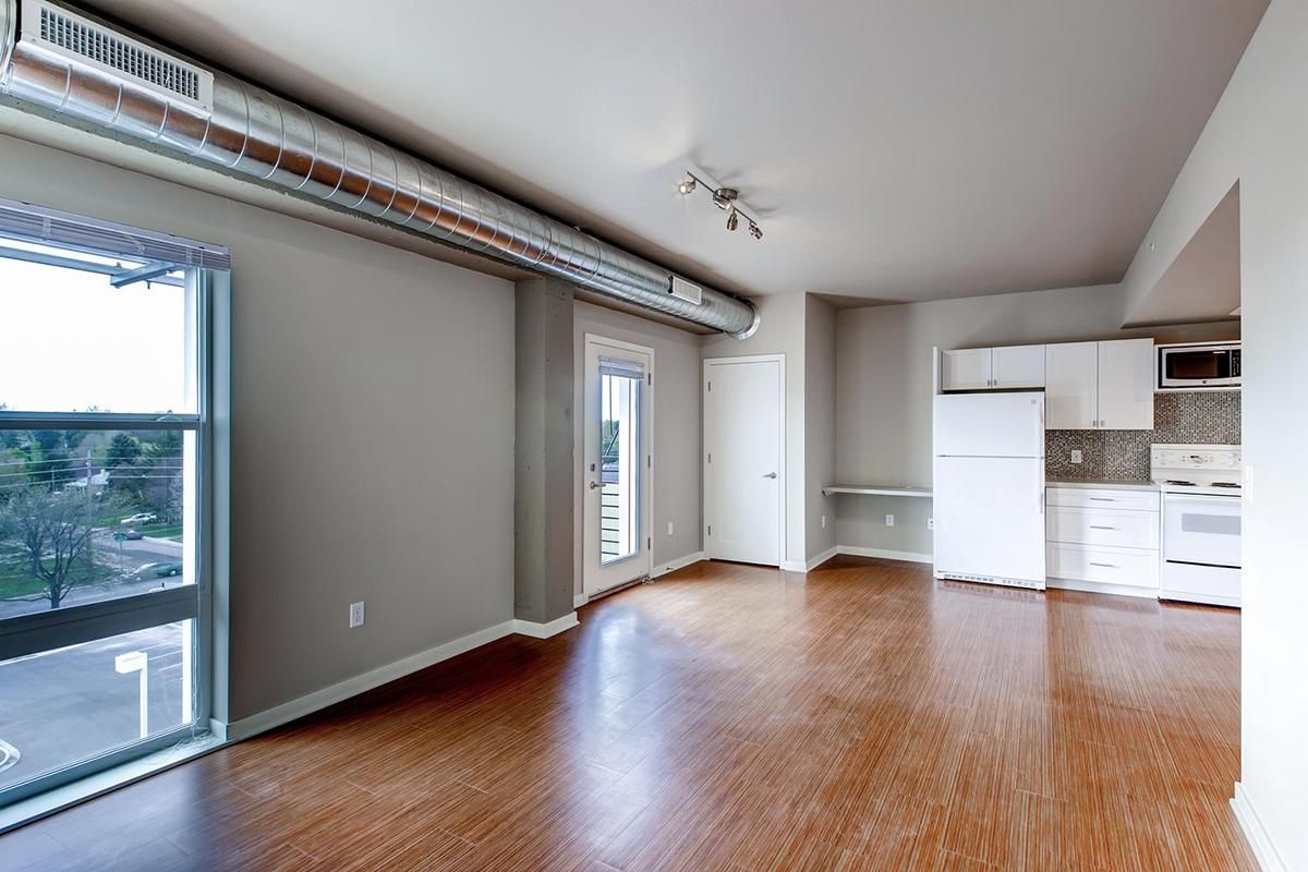 Empty apartment interior with wooden floor, large windows, and white kitchen appliances.