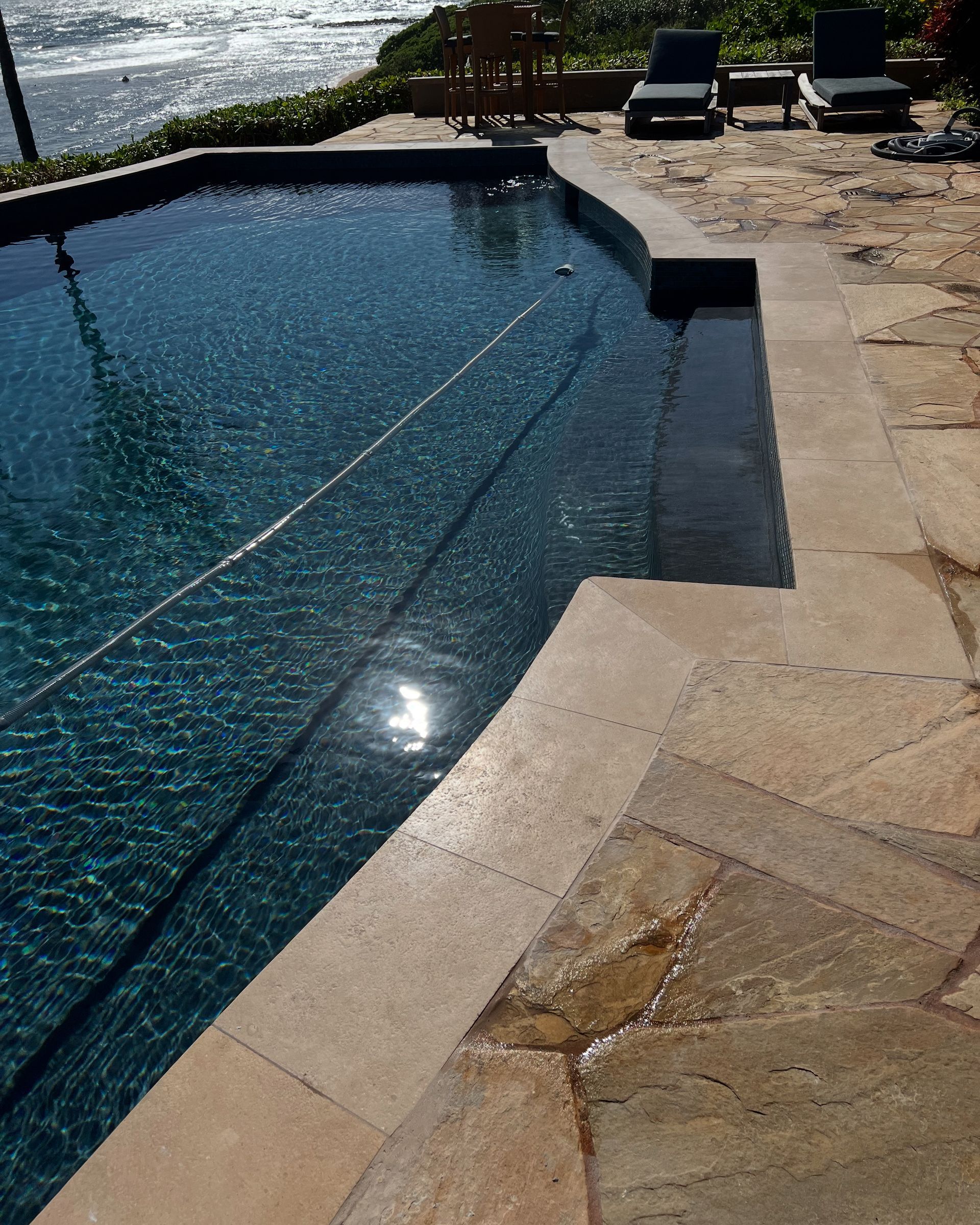 Pool with steps, tan stone patio, and ocean in the background. Reflective sunlight on the water's surface.