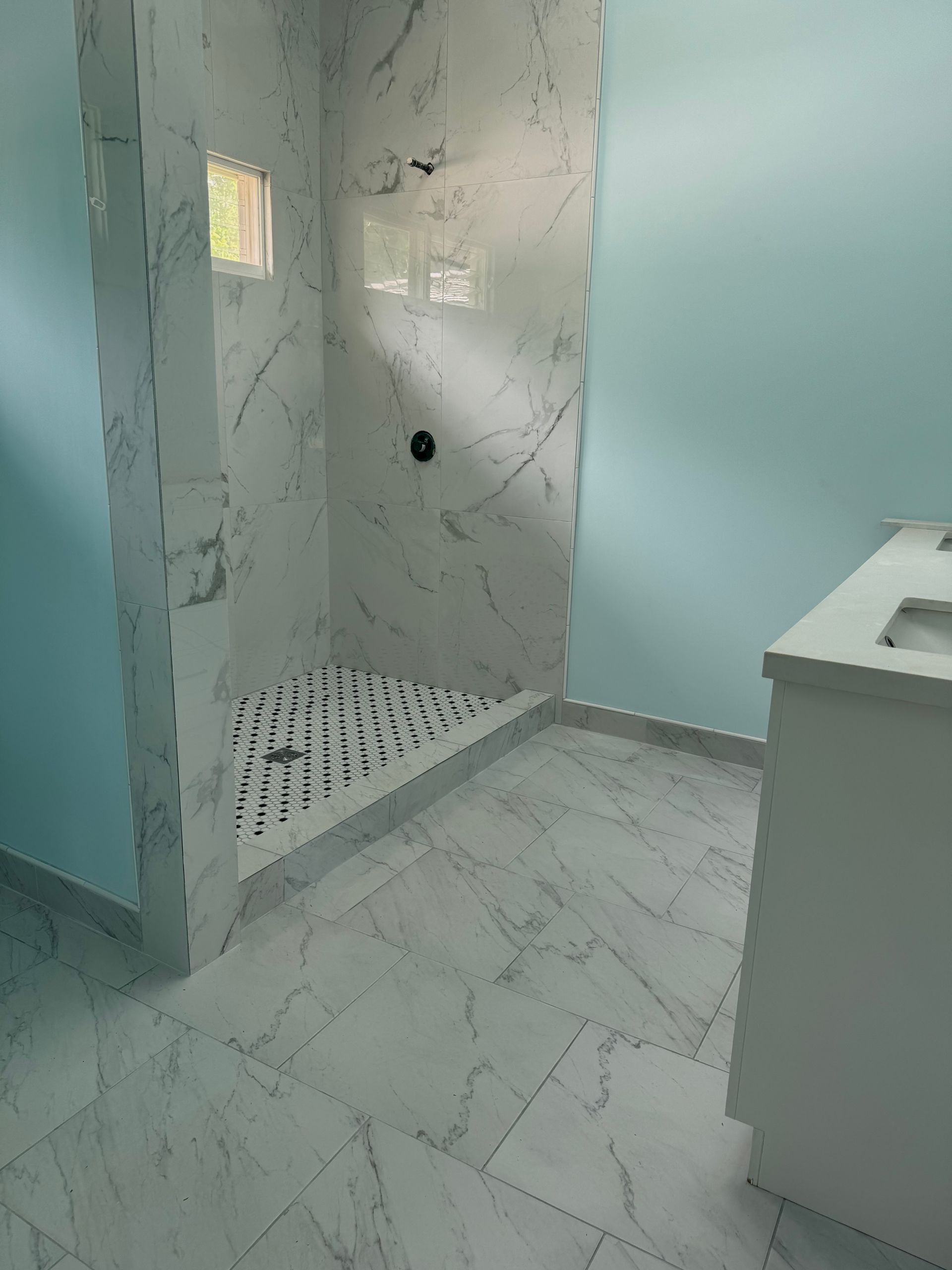 Bathroom with white marble-like tile. Shower with black fixtures and patterned floor. Light blue walls, white cabinet.