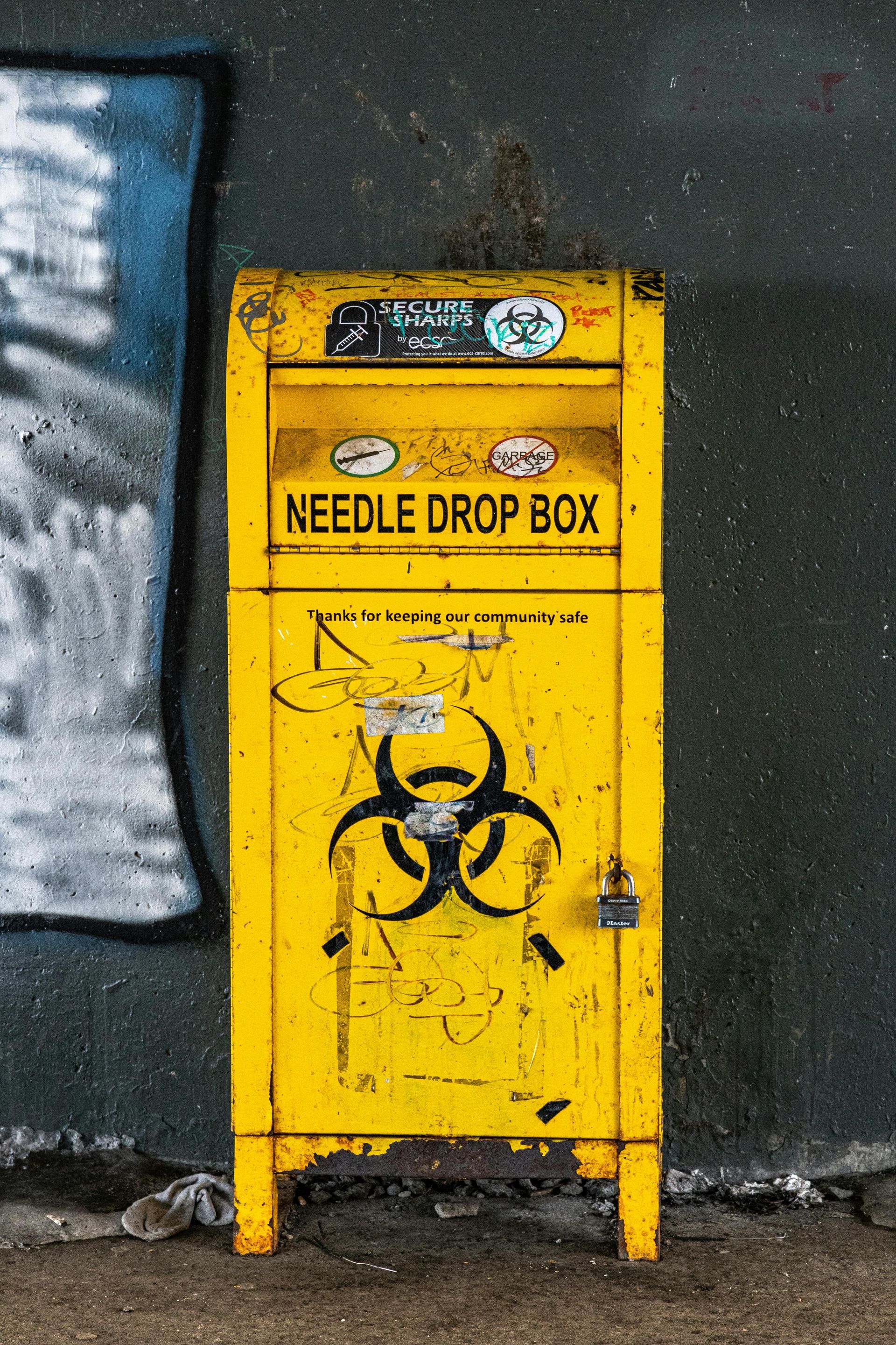 Three old televisions and a mattress discarded on a grassy area, next to a brick wall with house numbers 58 and 60.