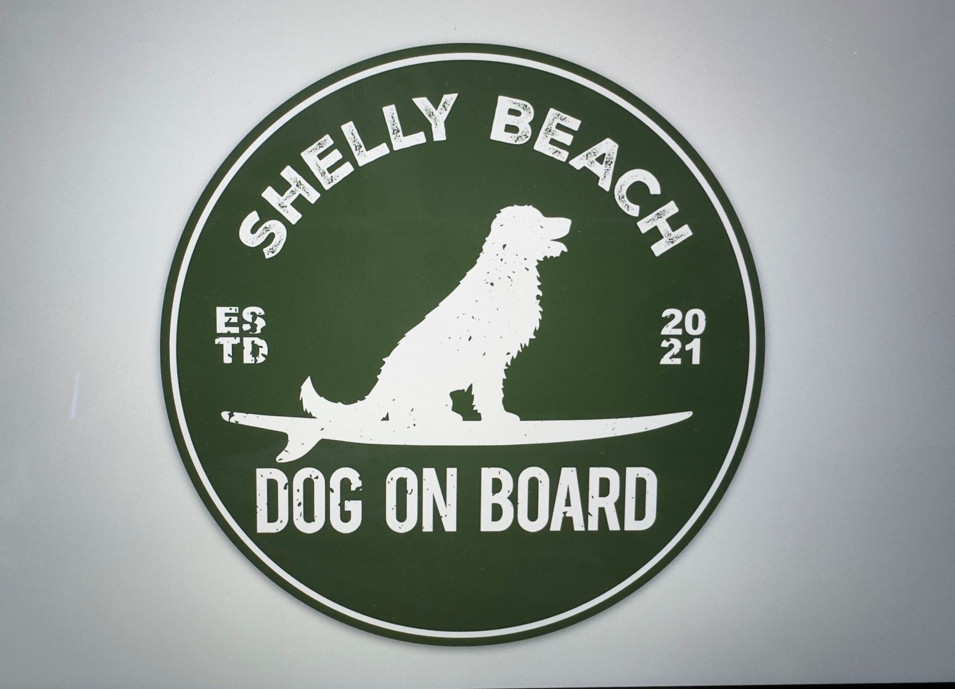 Shelly Beach logo. Green circle with white text and dog surfing on a board. 