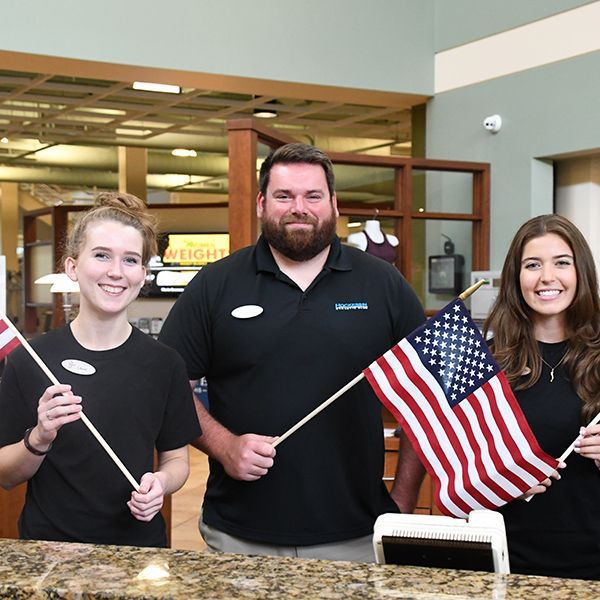 Three members of Hockessin Athletic Club's Front Desk team smiling and waving American flags