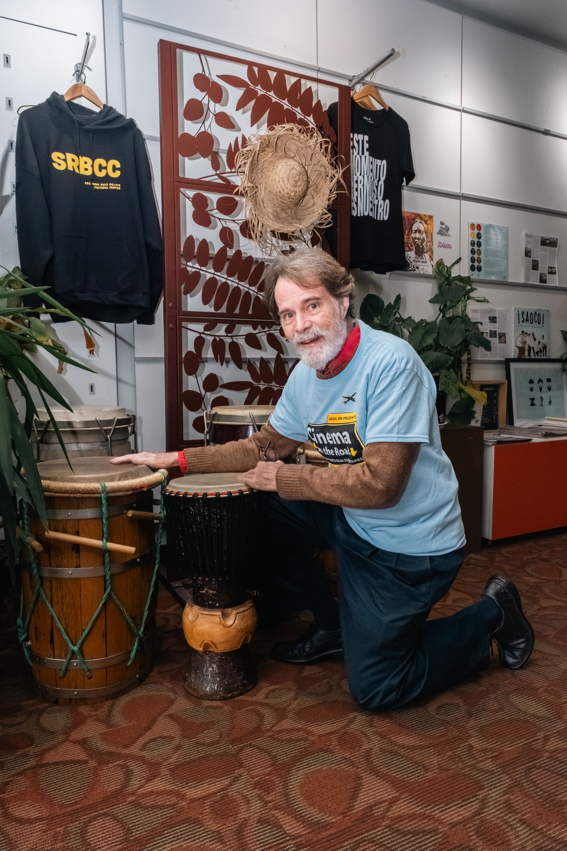 Man kneeling, pointing at drums; shop interior with merchandise and a plant. Portrait for events
