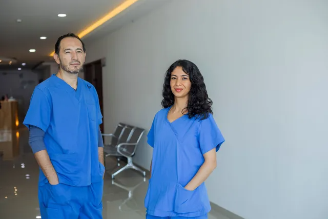 Two medical professionals in blue scrubs stand in a hallway, smiling at the camera.