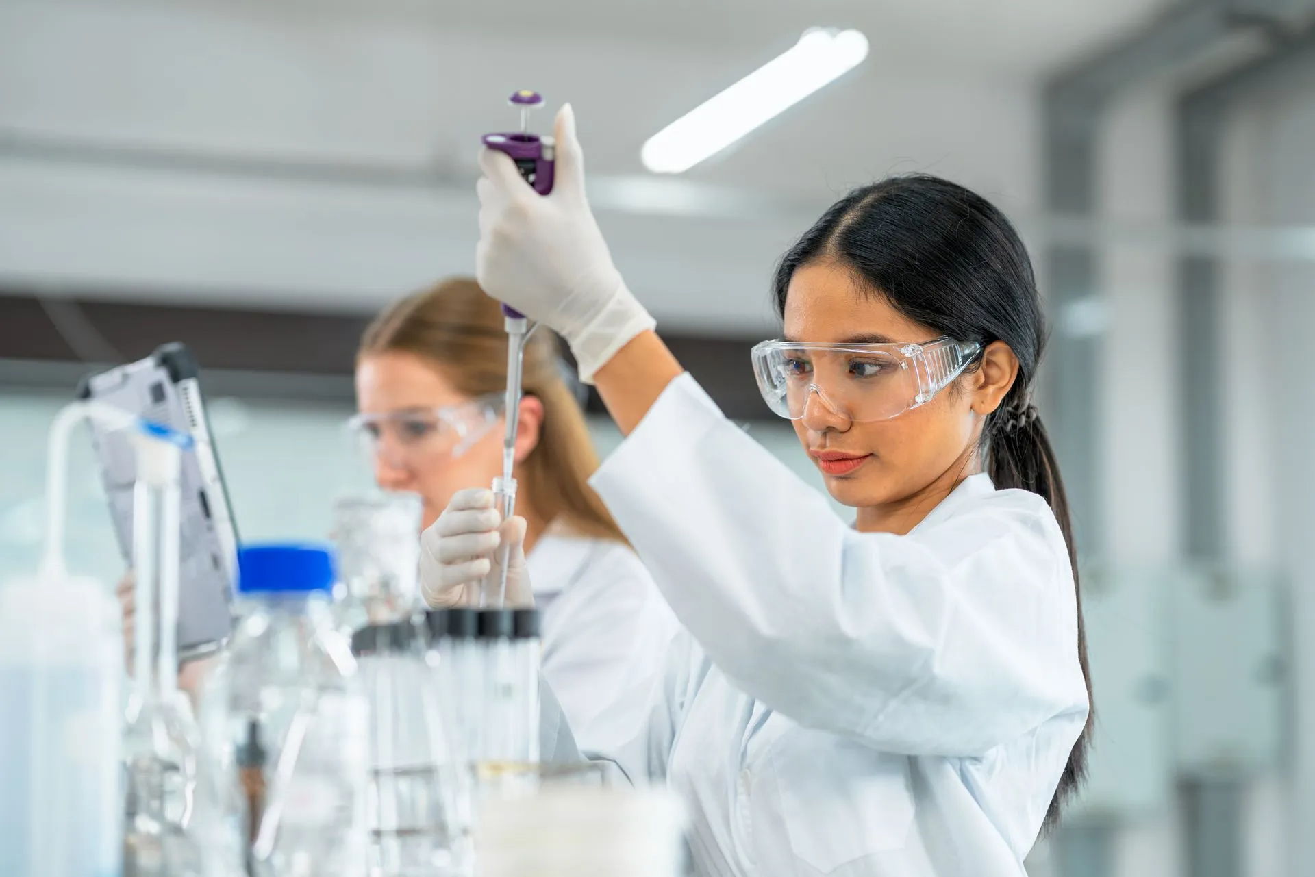 Woman in lab coat and goggles using pipette in a lab setting.
