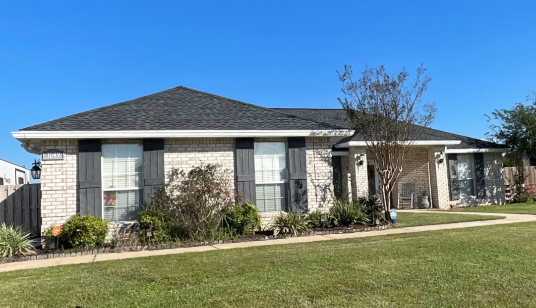 A brick house with a gray roof and black shutters is sitting on top of a lush green lawn.