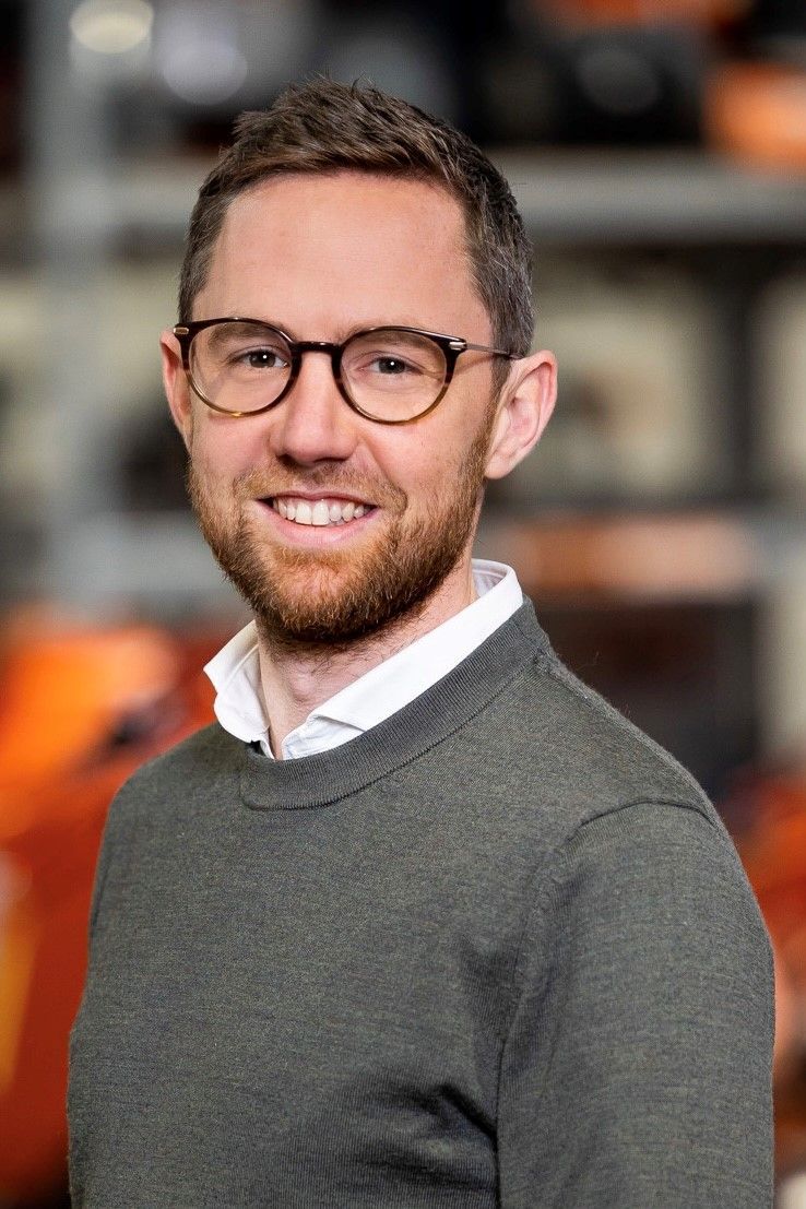 Een man met een bril en een baard, gekleed in een grijze trui, staat lachend in een fabrieksomgeving.