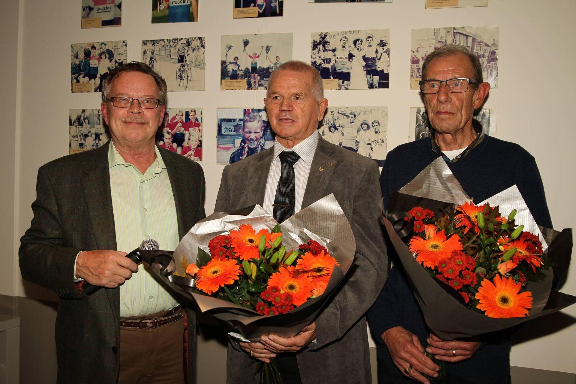 Drie mannen met boeketten staan ​​lachend voor ingelijste foto's.
