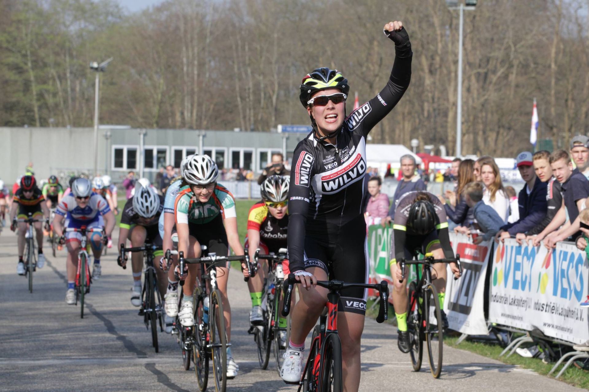 Een wielrenner steekt triomfantelijk zijn vuist in de lucht bij de finish, aan de leiding van een groep renners.