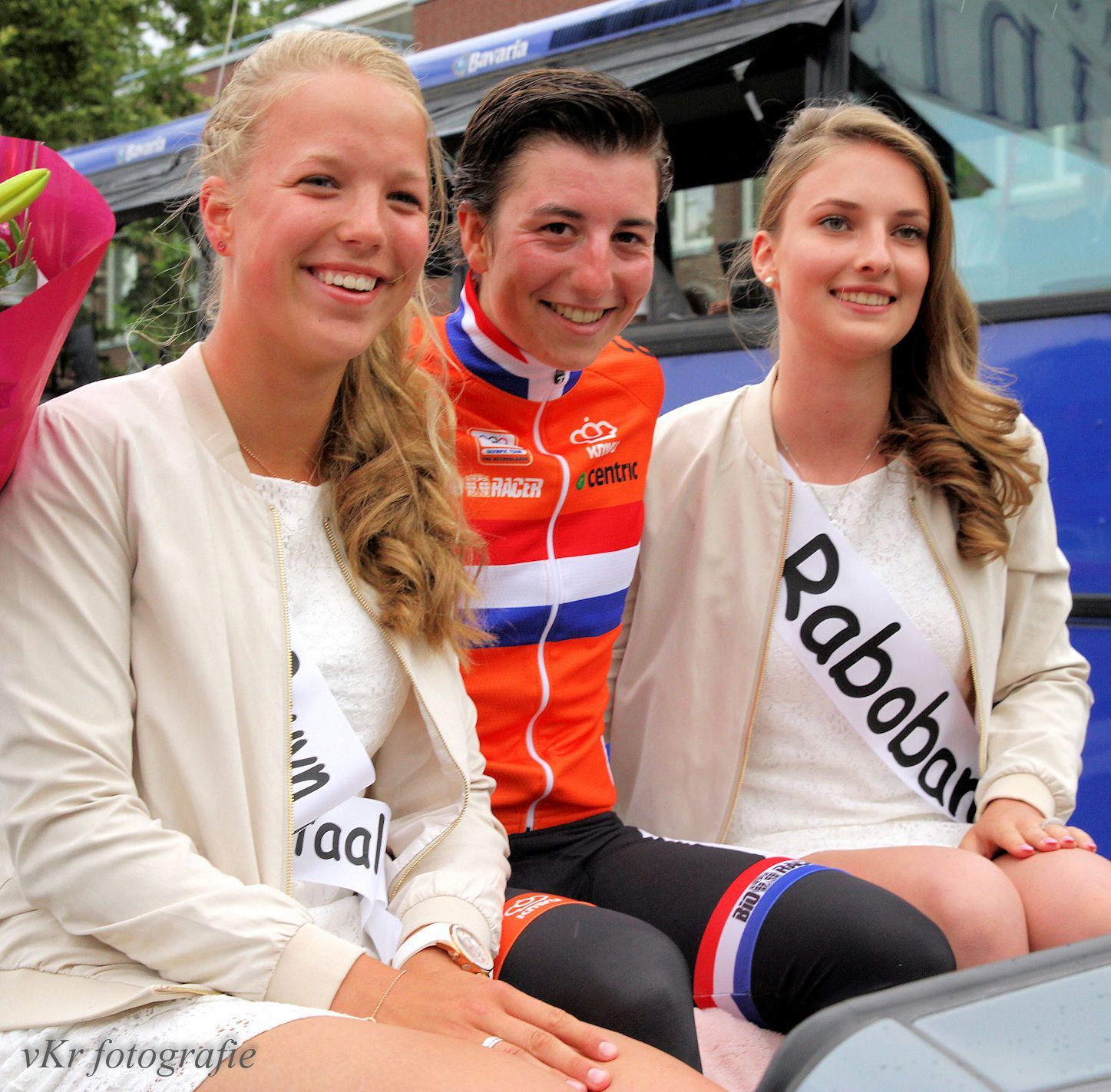 Een fietser in oranje uniform lacht naar twee vrouwen met sjerpen, die op een voertuig zitten.