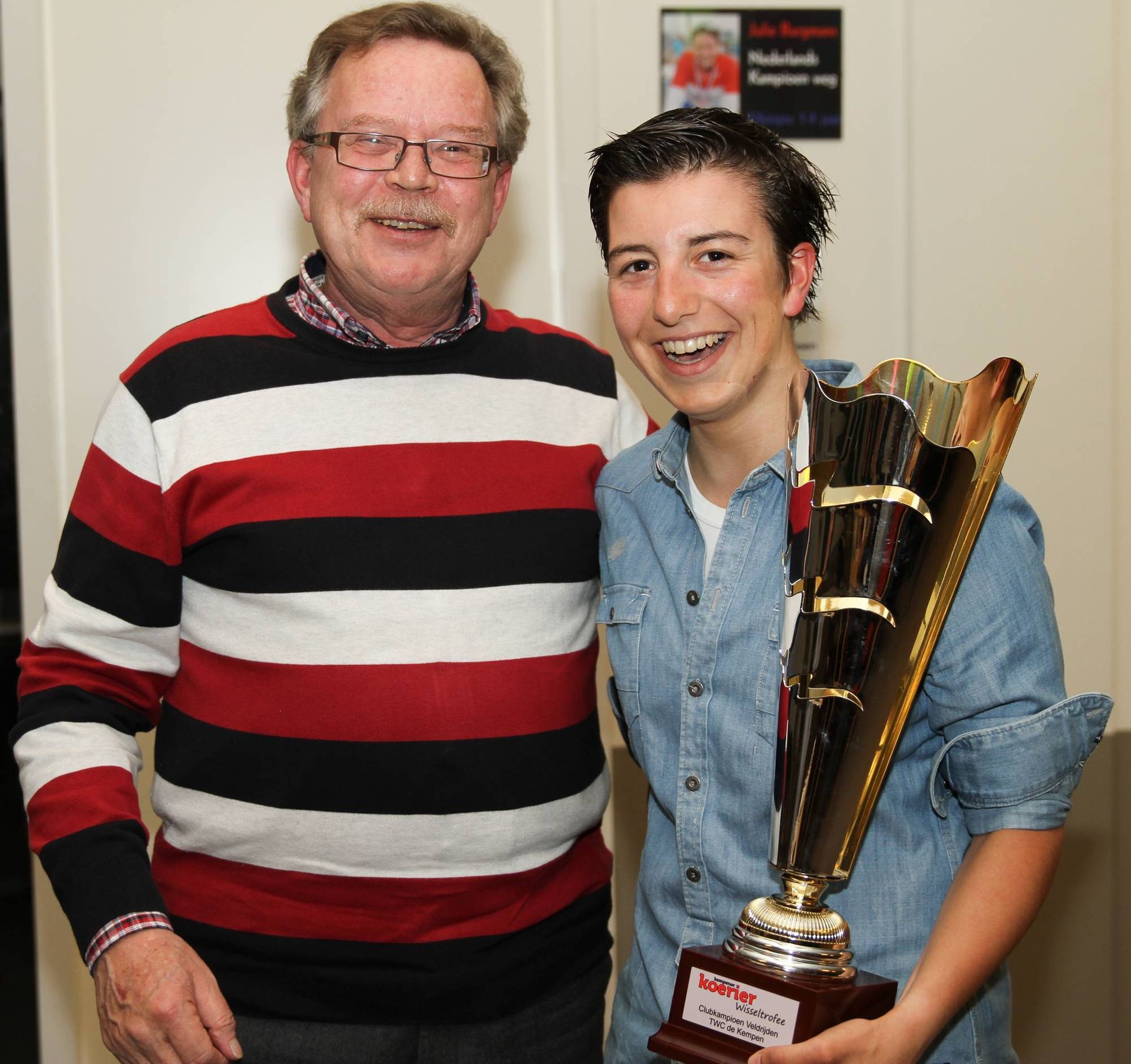 Een man en een jongere glimlachen en houden een trofee vast. De man draagt ​​een bril en een rood-zwarte trui. 