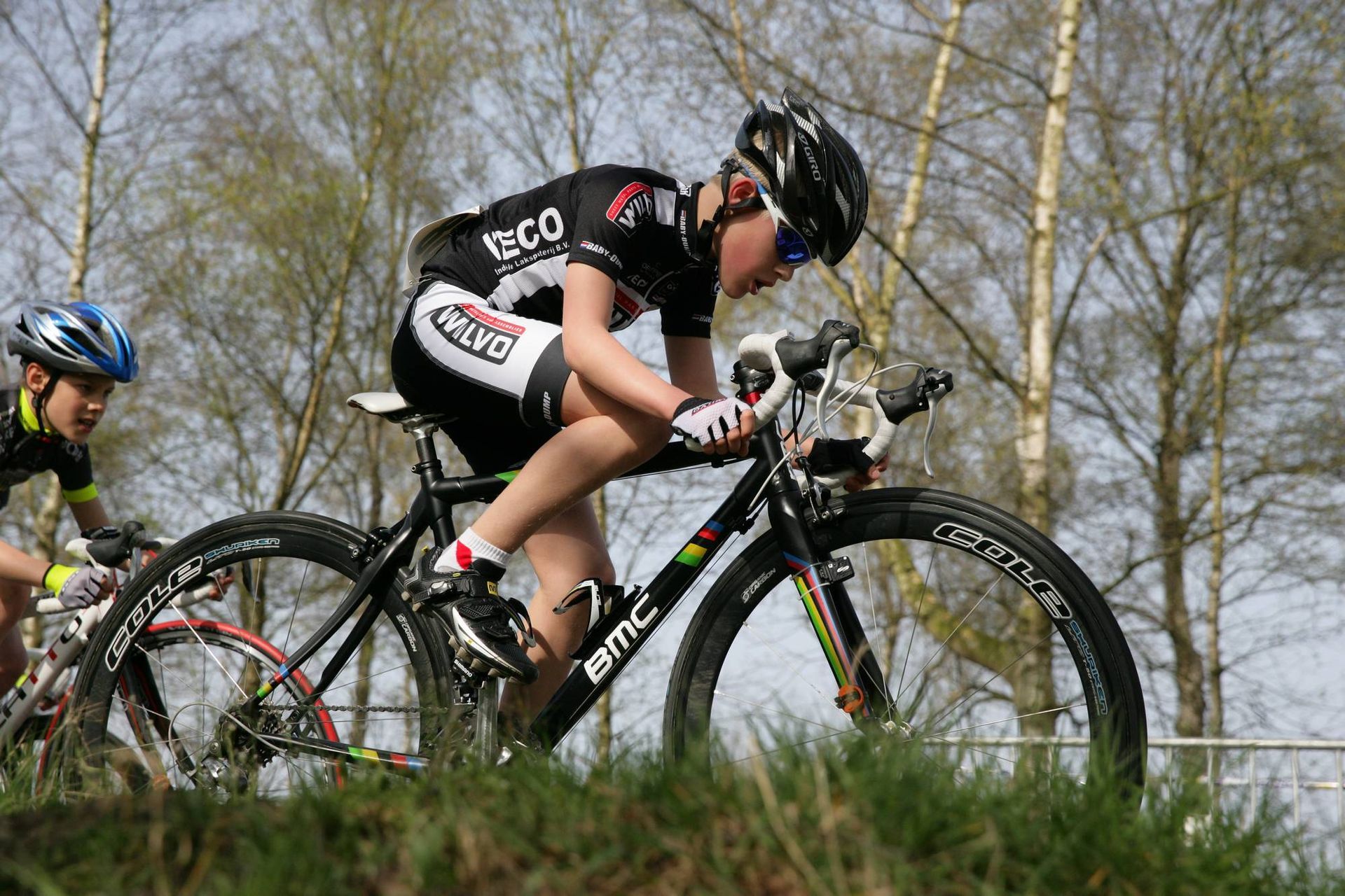 Een fietser op een zwarte fiets rijdt over het gras, gekleed in een zwart-wit tenue en helm. Een andere fietser volgt.