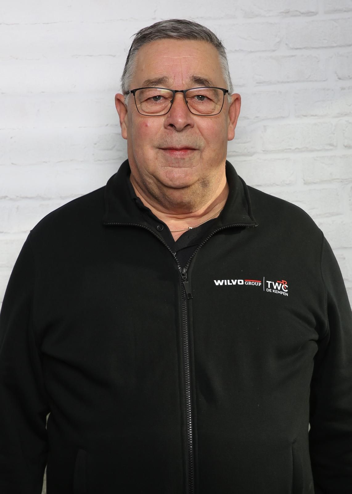 Man met bril en zwarte jas met rits tegen een witte bakstenen muur. De jas heeft witte tekst en een rood logo.