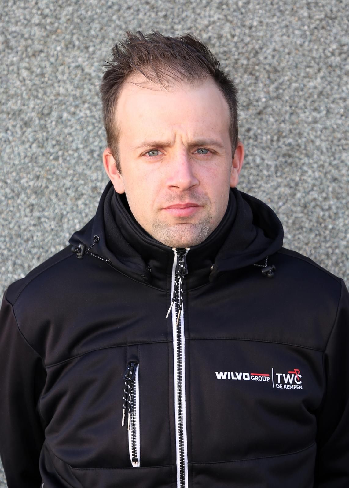 Een man in een zwarte jas met capuchon kijkt in de camera tegen een grijze muur.