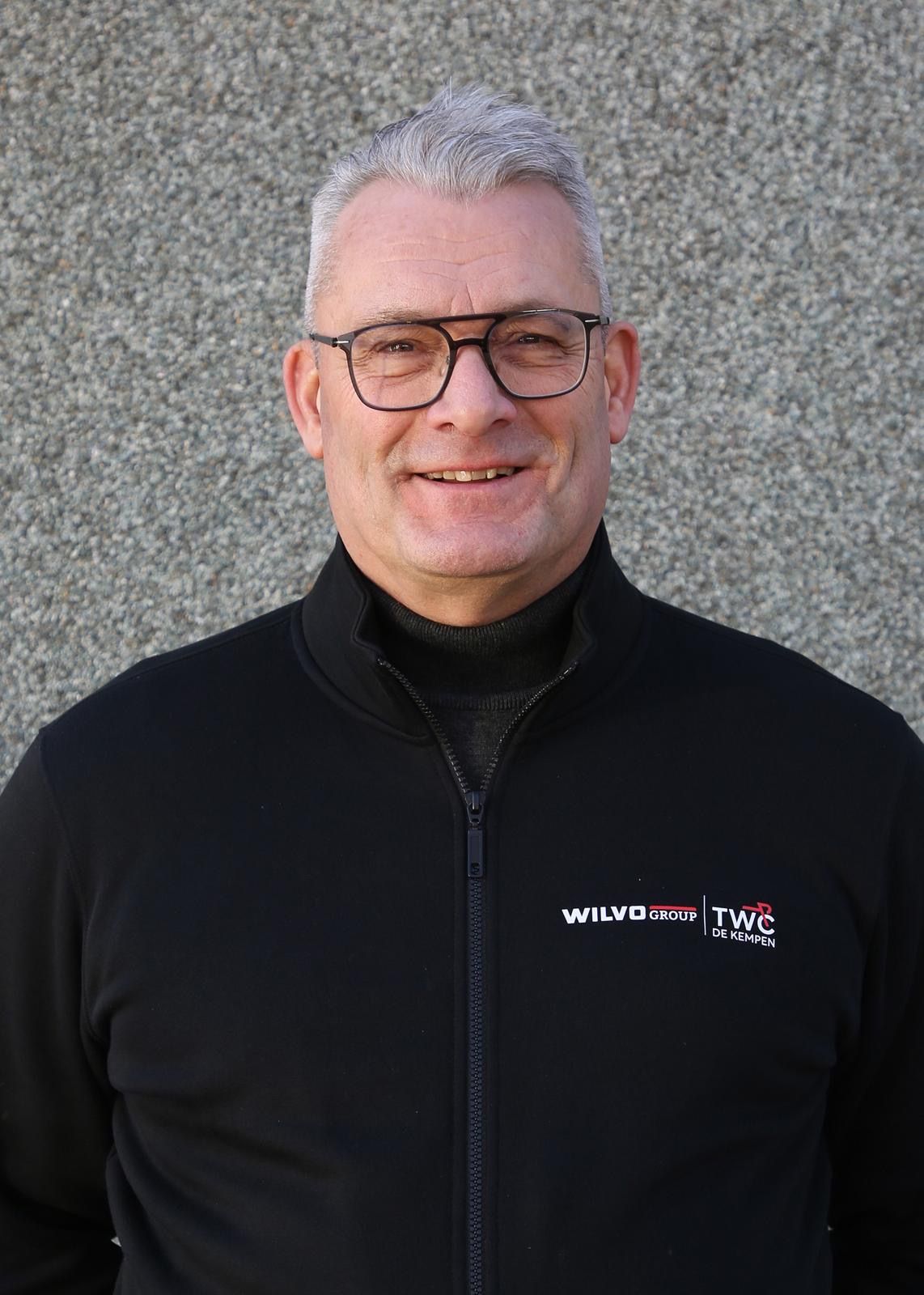 Man met bril, zwarte jas aan, lachend. Grijs haar, staat buiten.