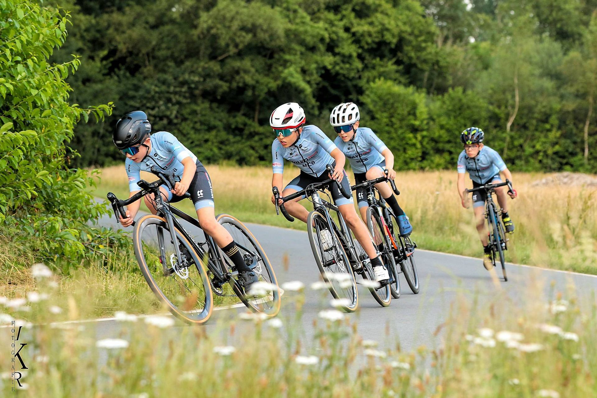 Wielrenners in lichtblauwe shirts racen op een geplaveide weg, in een landelijke omgeving met wilde bloemen.